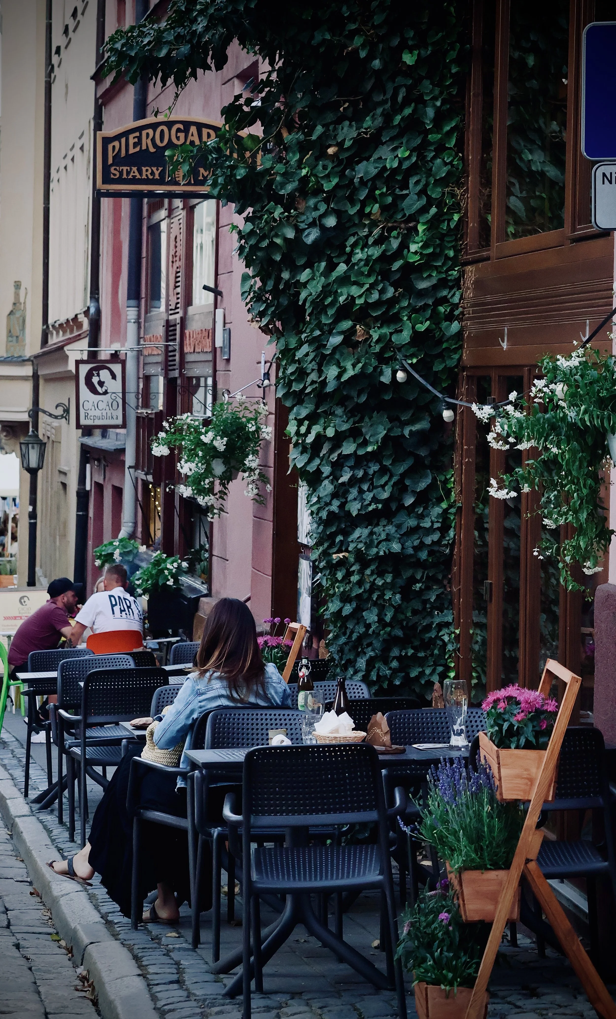 Outdoor cafe with black chairs and tables, some with glasses and flowers, set along a cobblestone street. People are seated and conversing. There is greenery and potted plants on the right. Buildings with signs and plants are visible in the backgroun