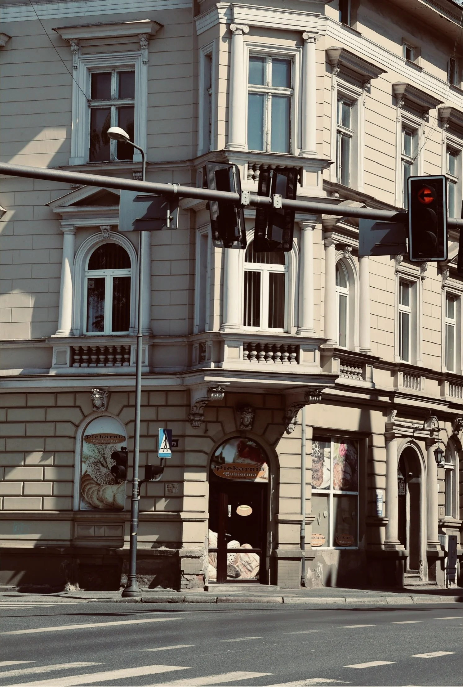 An ornate beige-colored building with decorative window trims and columns, situated at a street corner. Signs indicate a bakery or pastry shop called 'Piekarnia Cukiernia'. There are traffic lights and street signs in the foreground, with a sidewalk 