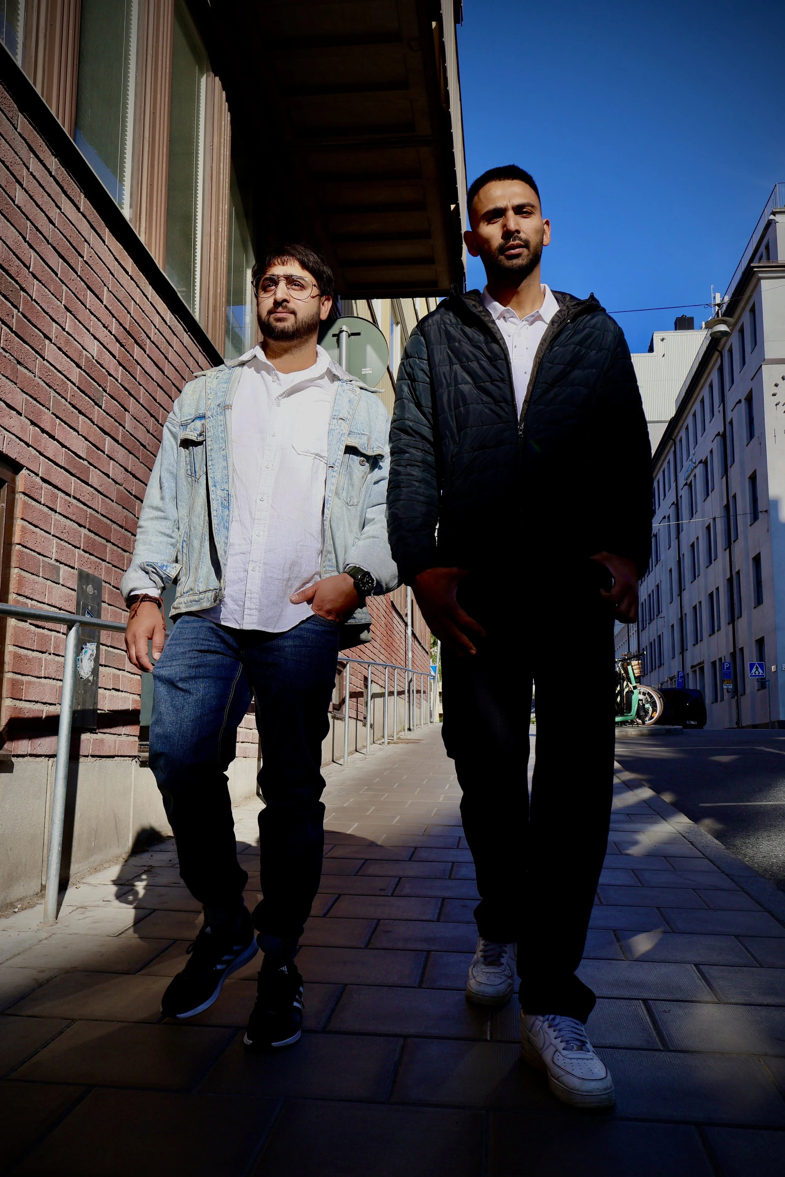 Two young men walking on a city sidewalk, with brick and white buildings in the background, both wearing casual clothing and sneakers, photographed during daytime under a bright blue sky.