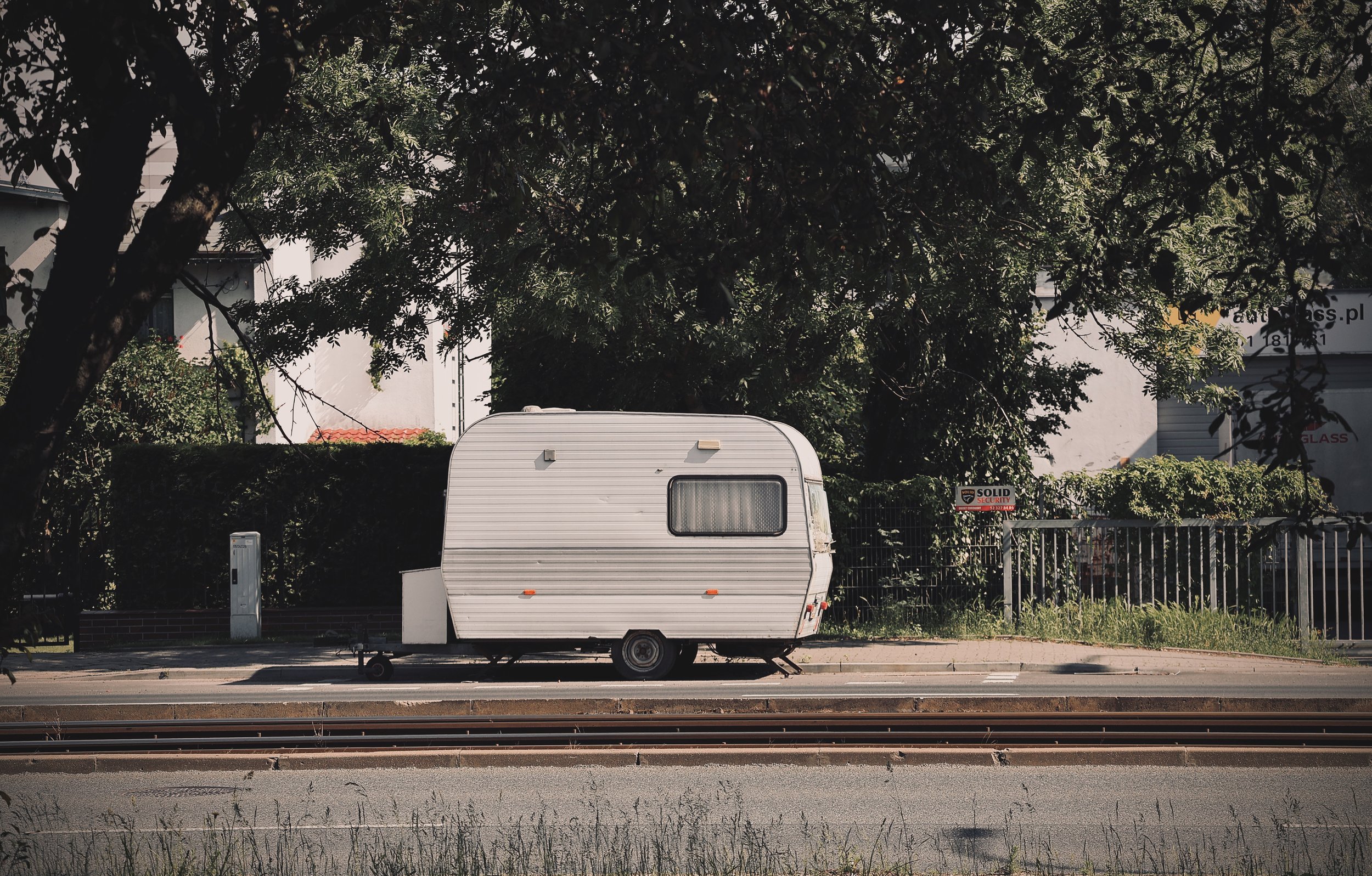 A white travel trailer parked on the side of a street in front of a fence with a sign that reads 'Solid Security'. There are large trees with dense foliage behind the fence, and buildings can be seen in the background.