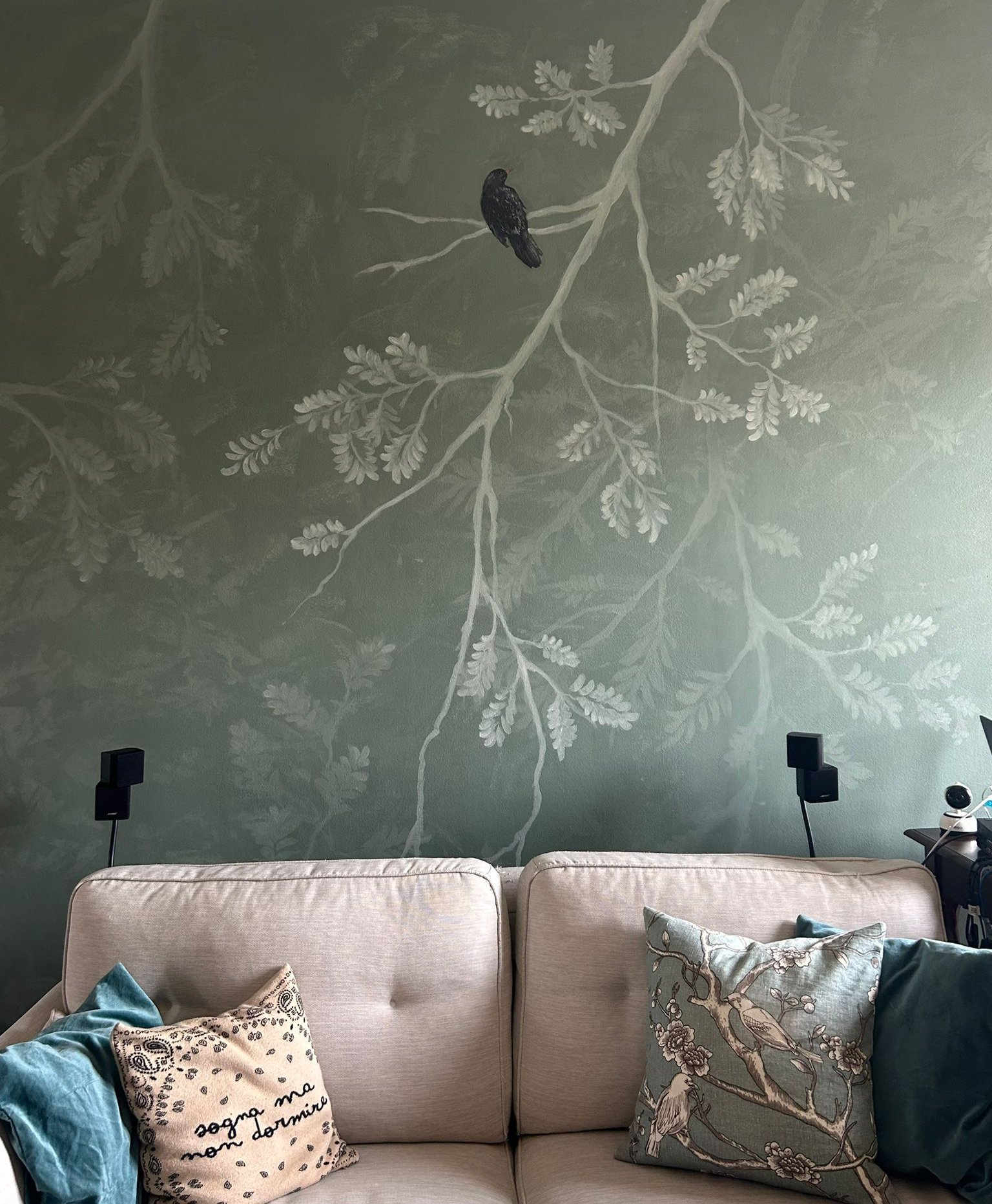 Soggiorno con divano beige decorato con cuscini, uno con scritte e altri con motivi di uccelli e fiori; sullo sfondo una parete verde con dipinto di rami e un uccello.