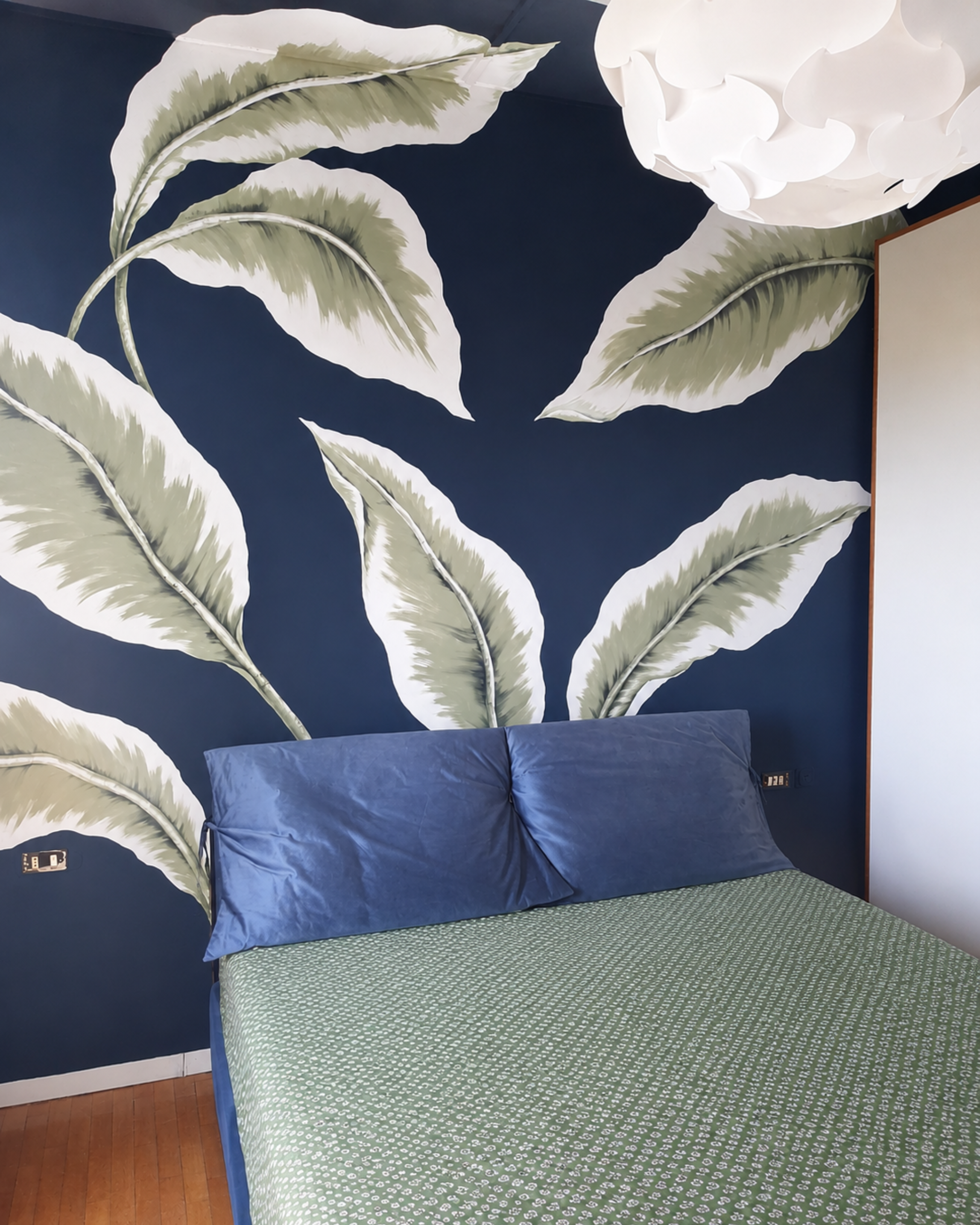 Letto con cuscini azzurri, sopra un comodino di plastica con presa elettrica, sfondo con carta da parati a foglie grandi verdi e bianche.