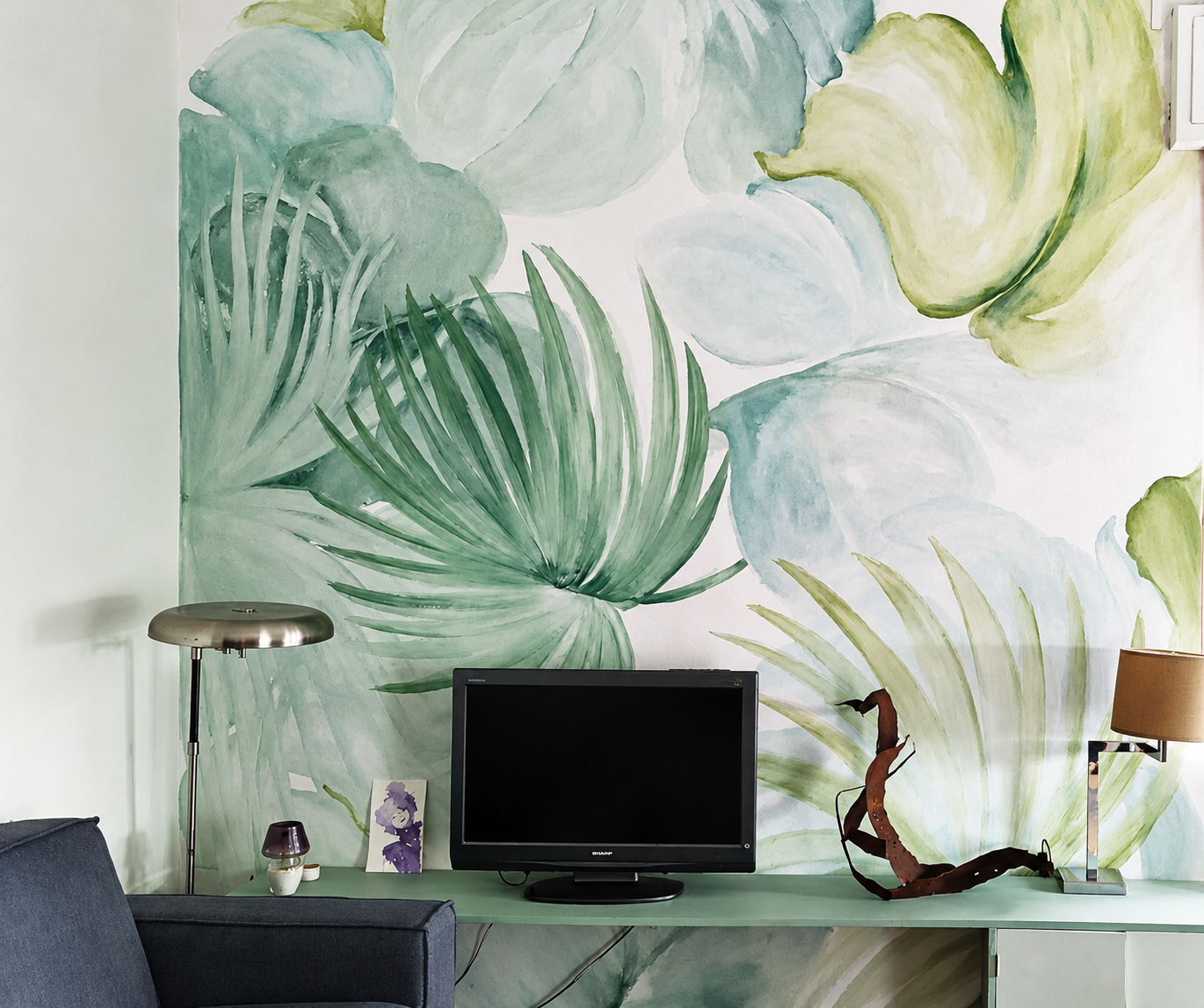 Muro con parete decorativa con grande stampa di foglie verdi su sfondo bianco, scrittoio verde con televisore, lampada da tavolo, lampada da terra e decorazioni