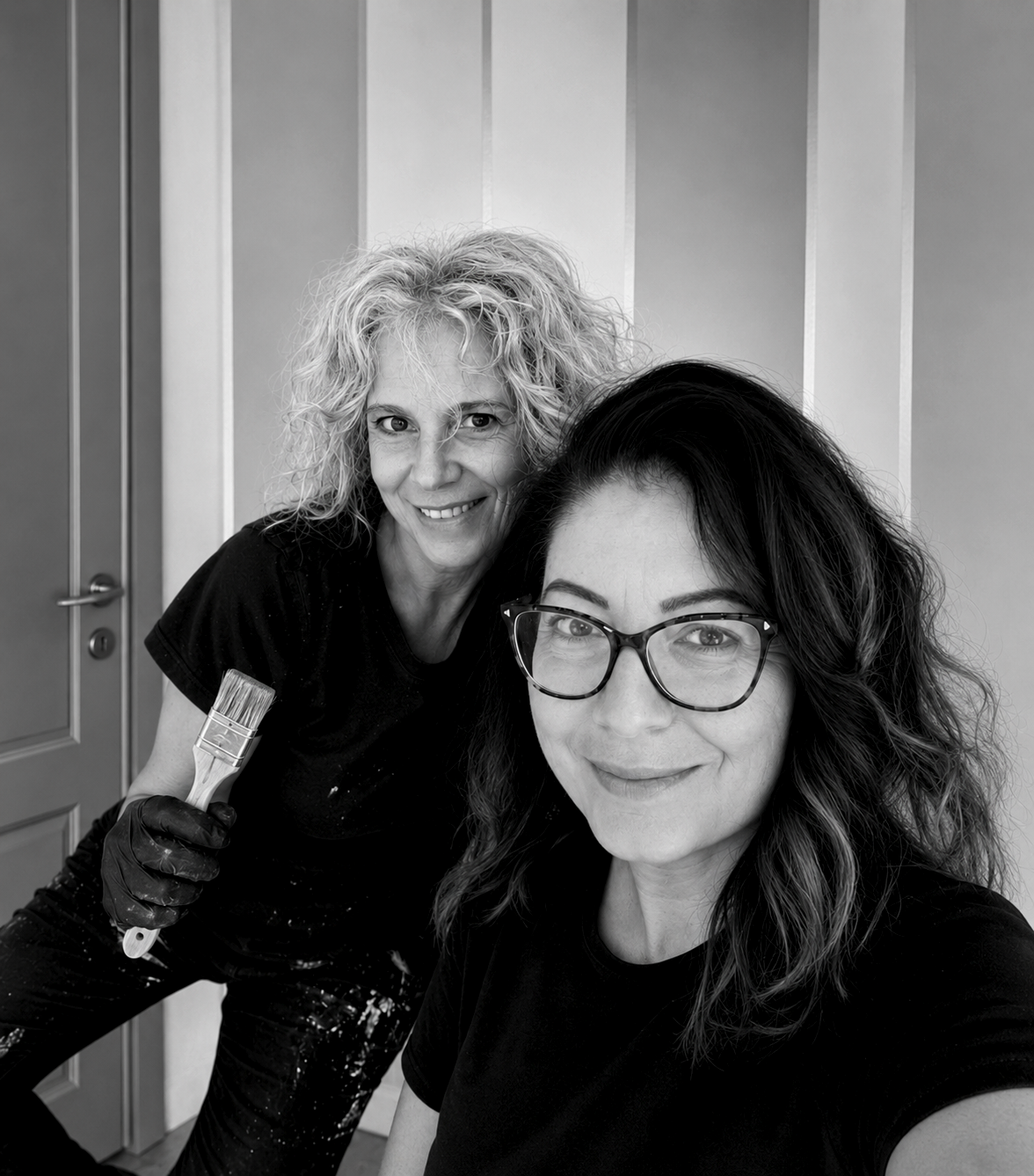 Due donne sorridenti, una con capelli ricci biondi e l'altra con capelli mossi castani e occhiali, mentre si preparano a dipingere una parete come professionisti della pittura.