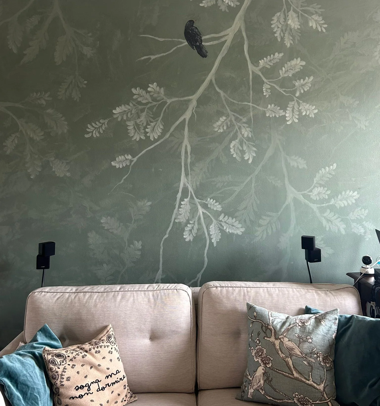Soggiorno con parete decorativa con mosaico di foglie e un uccello appoggiato su un ramo, divano beige con cuscini decorativi e diffusori acustici.