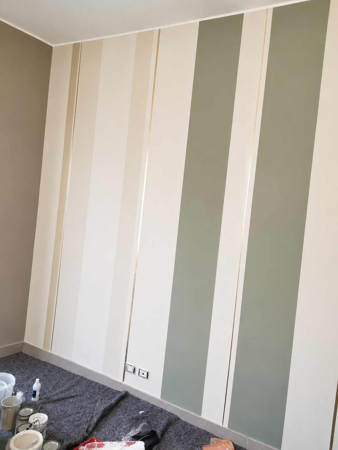 Muro con strisce verticali di colori beige, bianco e verde salvia, in fase di decorazione o pittura, con attrezzi e materiali per la verniciatura sul pavimento.
