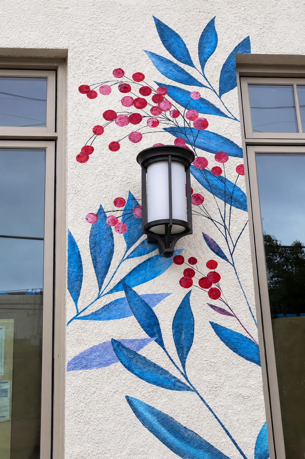 Murale decorativa con una lampada a parete e foglie blu e bacche rosse dipinte sul muro bianco vicino a due finestre.