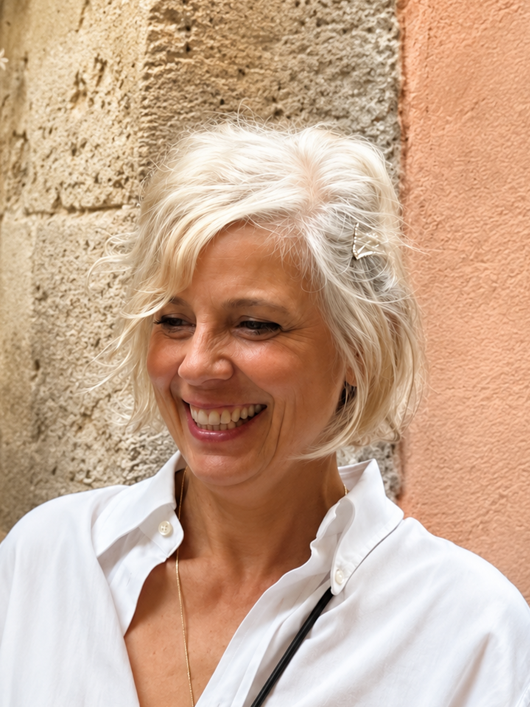 Donna sorridente con capelli biondi corti, indossa camicia bianca e ha uno stetoscopio. Sfondo con muro in pietra e parete rosa.