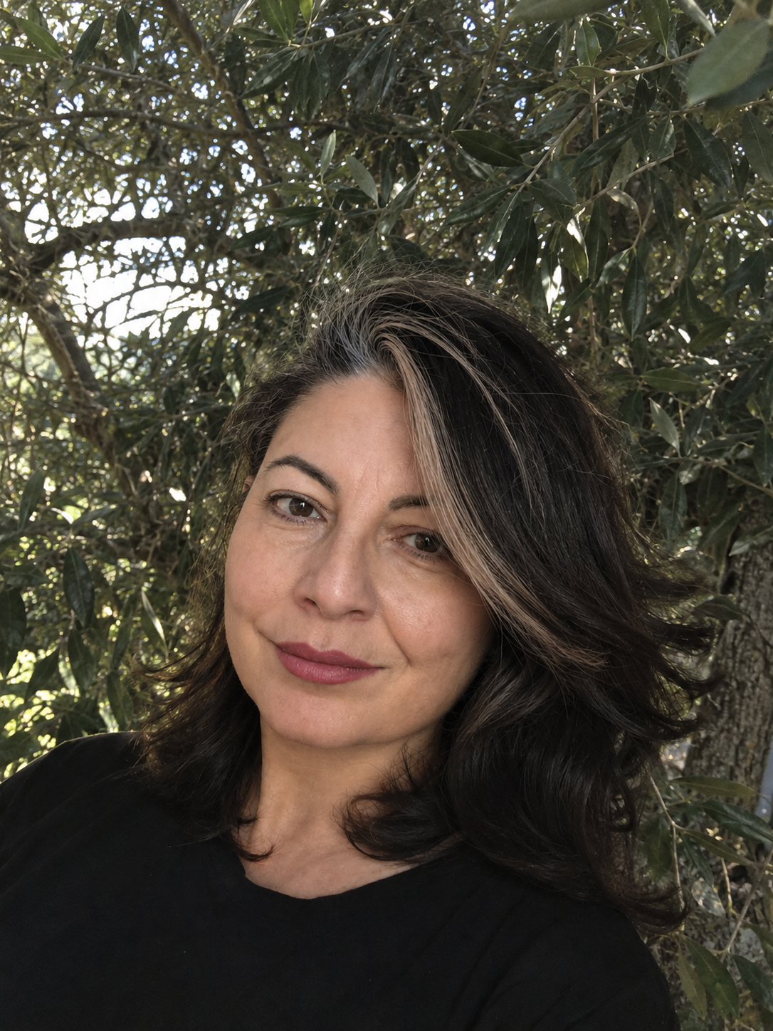 Una donna con capelli castani e bianchi che sorride, fotografata all'aperto davanti a foglie di un albero.