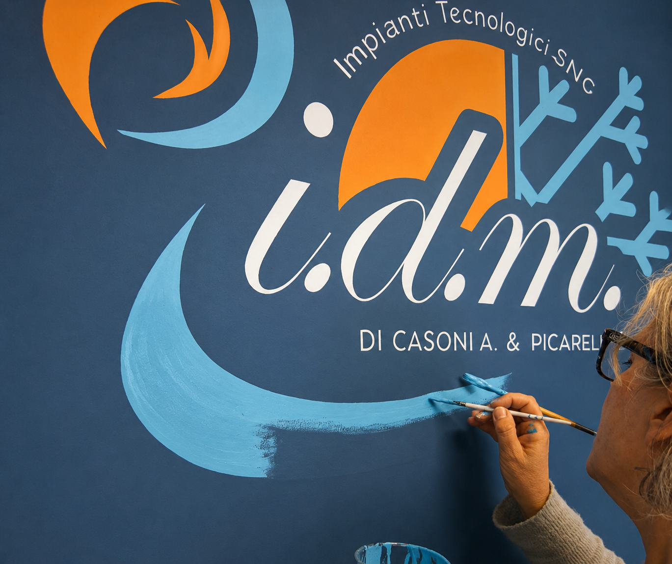 Una persona sta dipingendo una parete con un murale che include il testo ".d.m." e altre parole e disegni astratti, con colori blu, arancione e bianco.