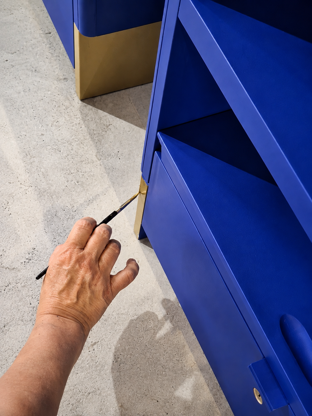 Una mano sta dipingendo una libreria blu con dettagli dorati usando un pennello.
