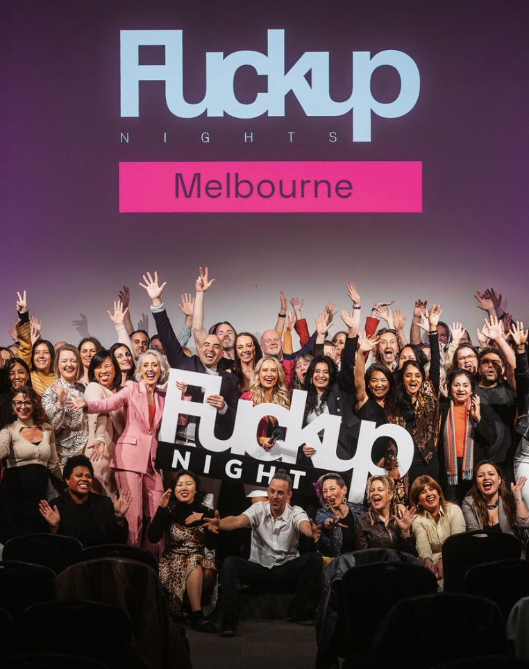 A large group of people on stage holding a sign that says 'F**kup Night' with a projector screen behind them displaying the same text and 'Melbourne'. The group appears happy and excited, with many raising their hands.