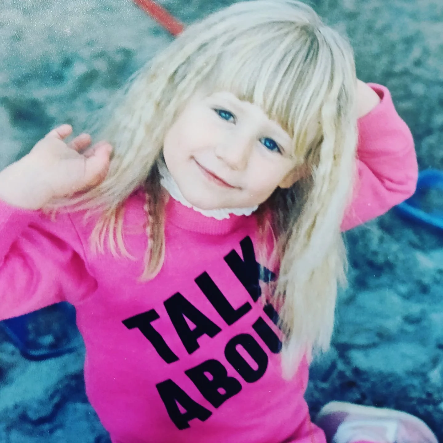 A young girl with blonde hair and blue eyes, smiling and wearing a pink sweatshirt with black text that says 'TALK ABOUT'. She is outdoors near water, holding her hair back with one hand.