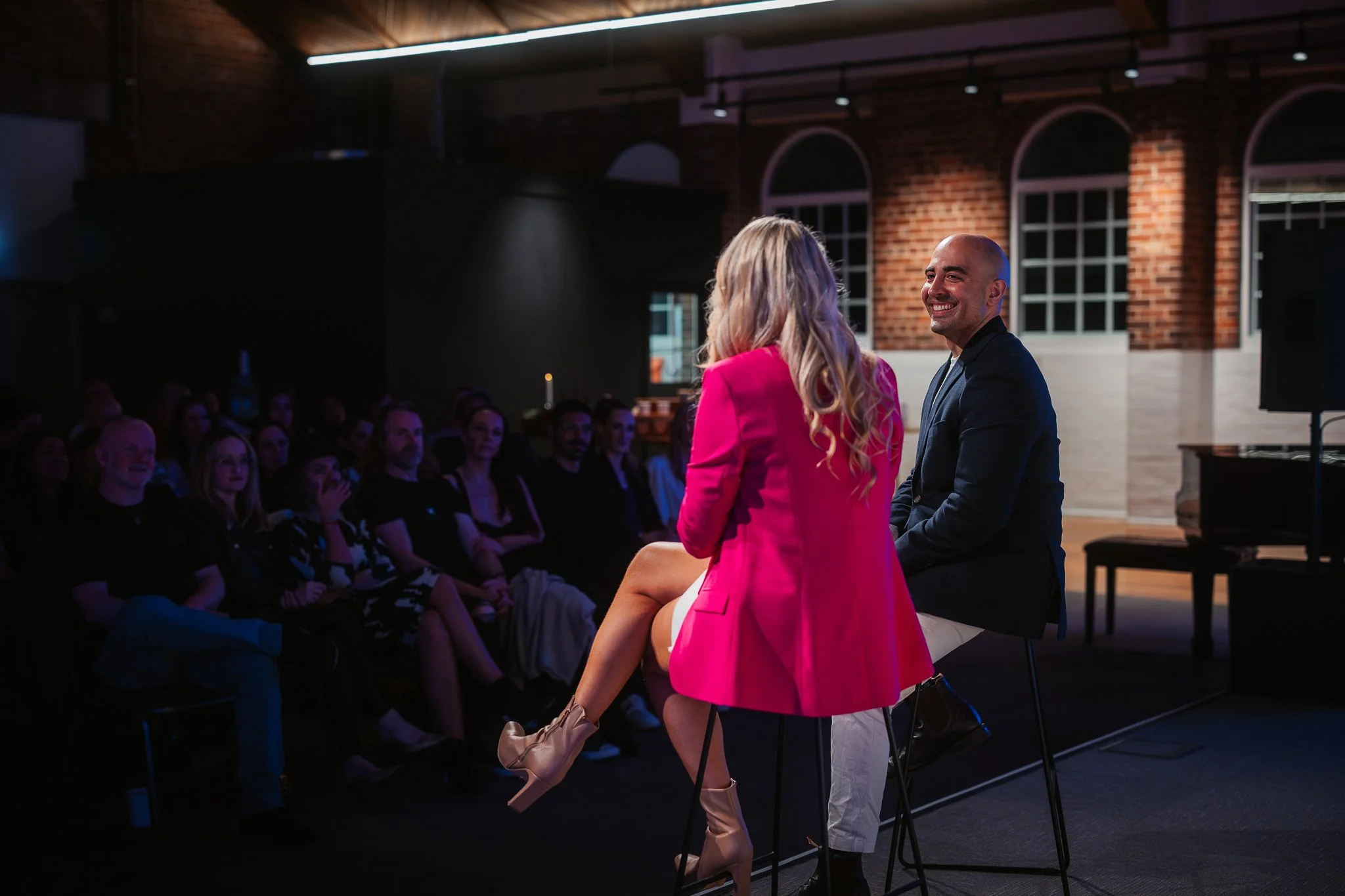 A woman in a pink blazer and beige ankle boots sitting on a tall black stool, talking to a man in a dark blazer and light pants, in front of an audience in a dimly lit venue with exposed brick walls and large windows.
