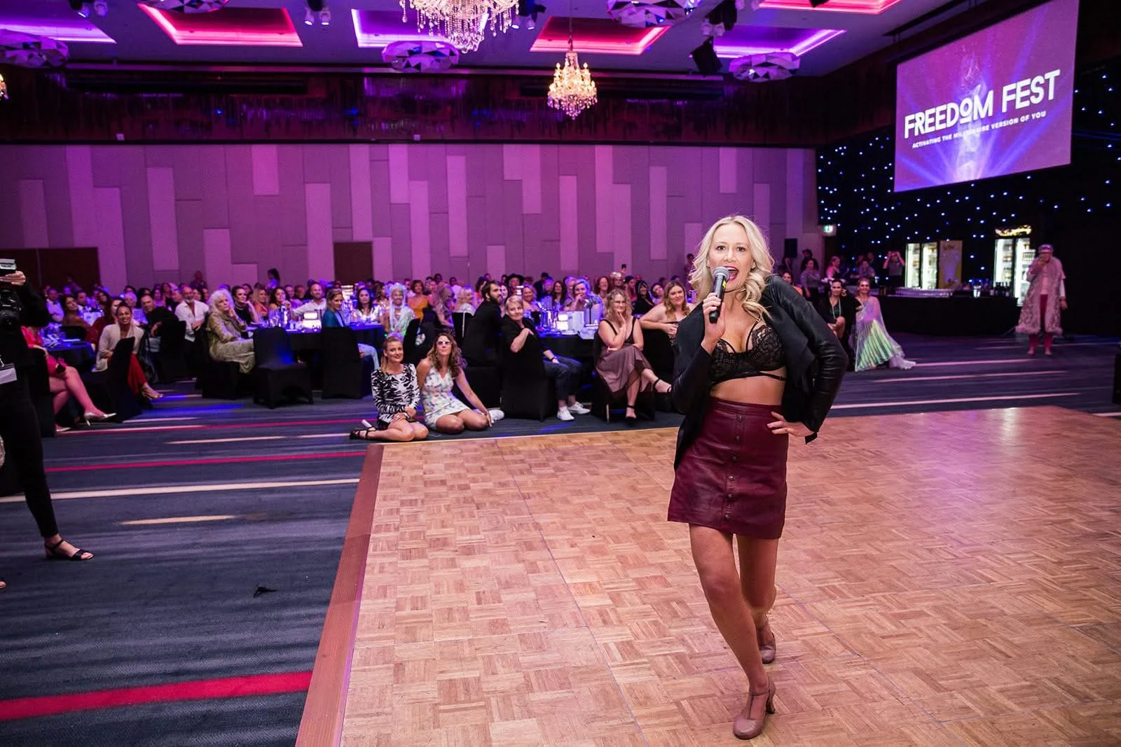 A woman with blonde hair, black lace top, maroon skirt, and high heels is singing into a microphone on a stage in front of a large audience in a decorated ballroom with chandeliers and a screen that says "FREEDOM FEST."