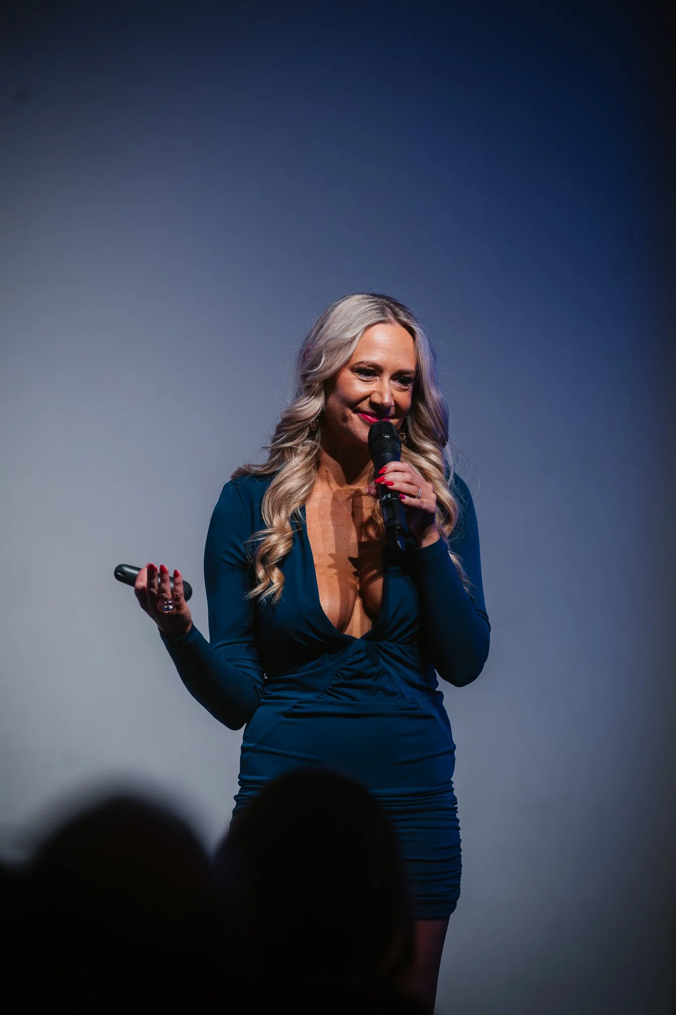 A woman with long, wavy blonde hair is standing on stage, holding a microphone in her right hand and a remote in her left hand. She is wearing a deep blue dress with a plunging neckline. The background is plain and dark.