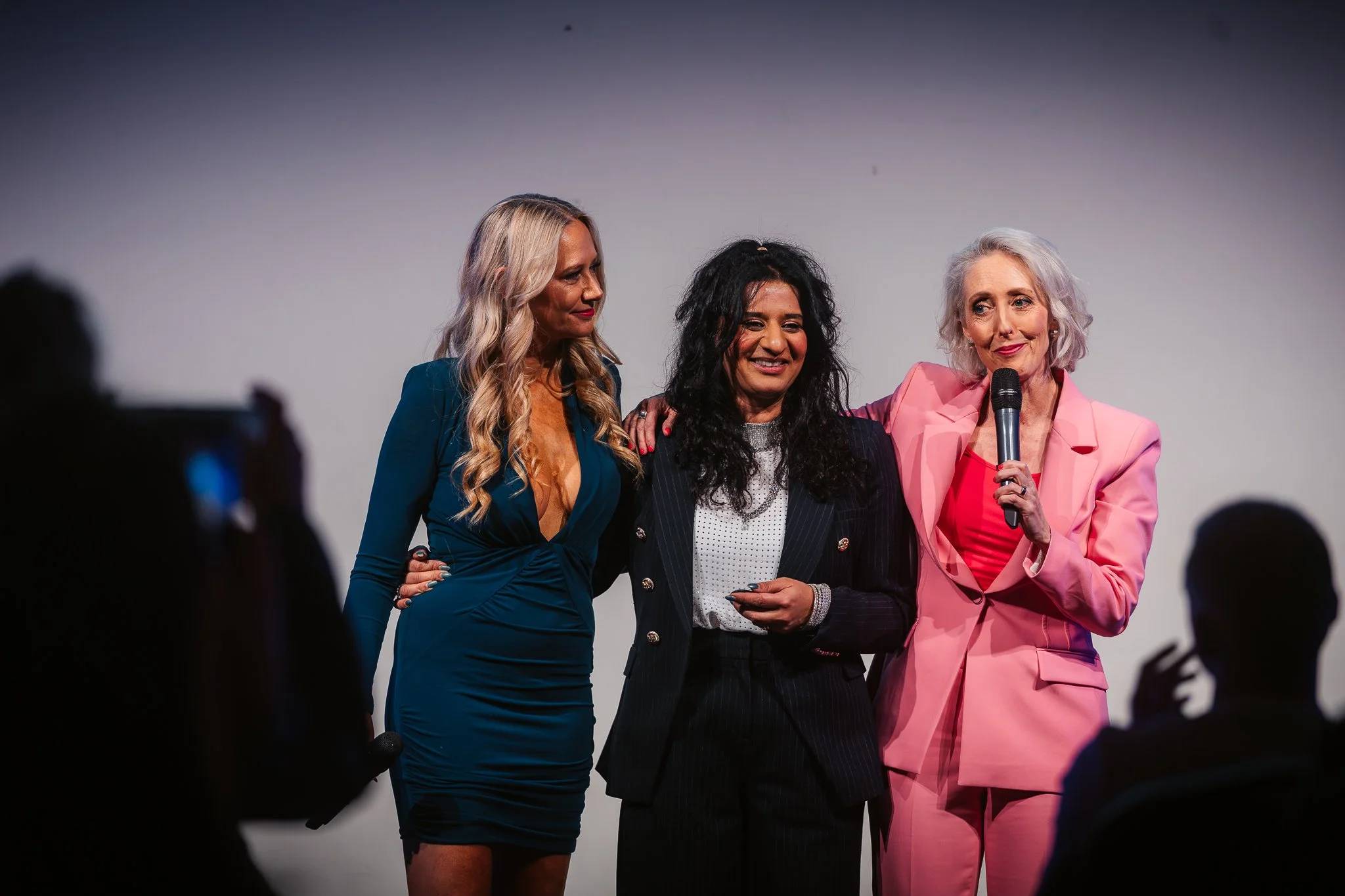 Three women standing on stage, with one woman holding a microphone, during an event. The woman on the left wears a teal dress, the woman in the middle wears a black pinstripe suit, and the woman on the right wears a pink suit. Audience members silhouette in foreground.