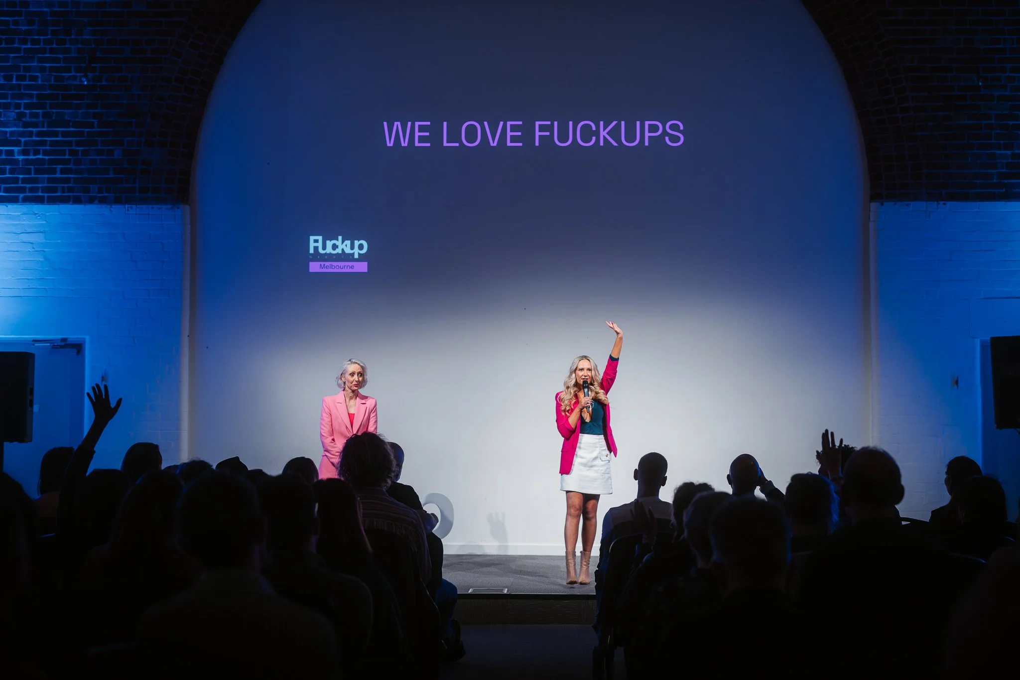 Women speaking on stage at a conference with a presentation that reads 'WE LOVE FUCKUPS' and 'Fuckup Melbourne'; audience watching, some with raised hands.