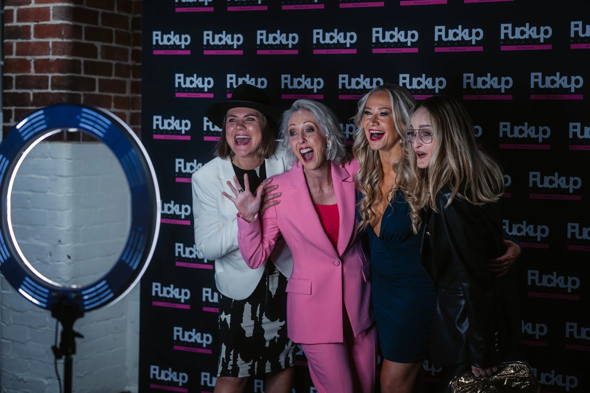 Four women posing together in front of a backdrop that reads 'Fuckup Melbourne'. They are smiling and appear to be enjoying themselves, dressed in fashionable clothing, with a ring light visible on the left side of the image.