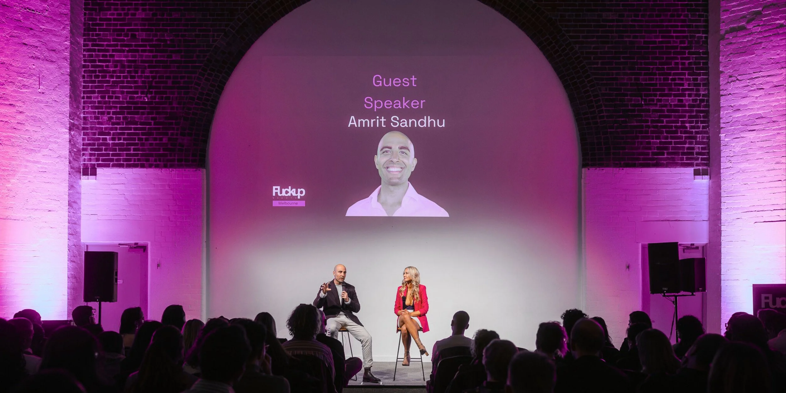 A conference stage with two speakers seated, a man and a woman, engaging in a discussion with an audience. A large screen behind them displays the name 'Amrit Sandhu' as a guest speaker, with his photo, and the event branding 'Flickp Melbourne'. The 