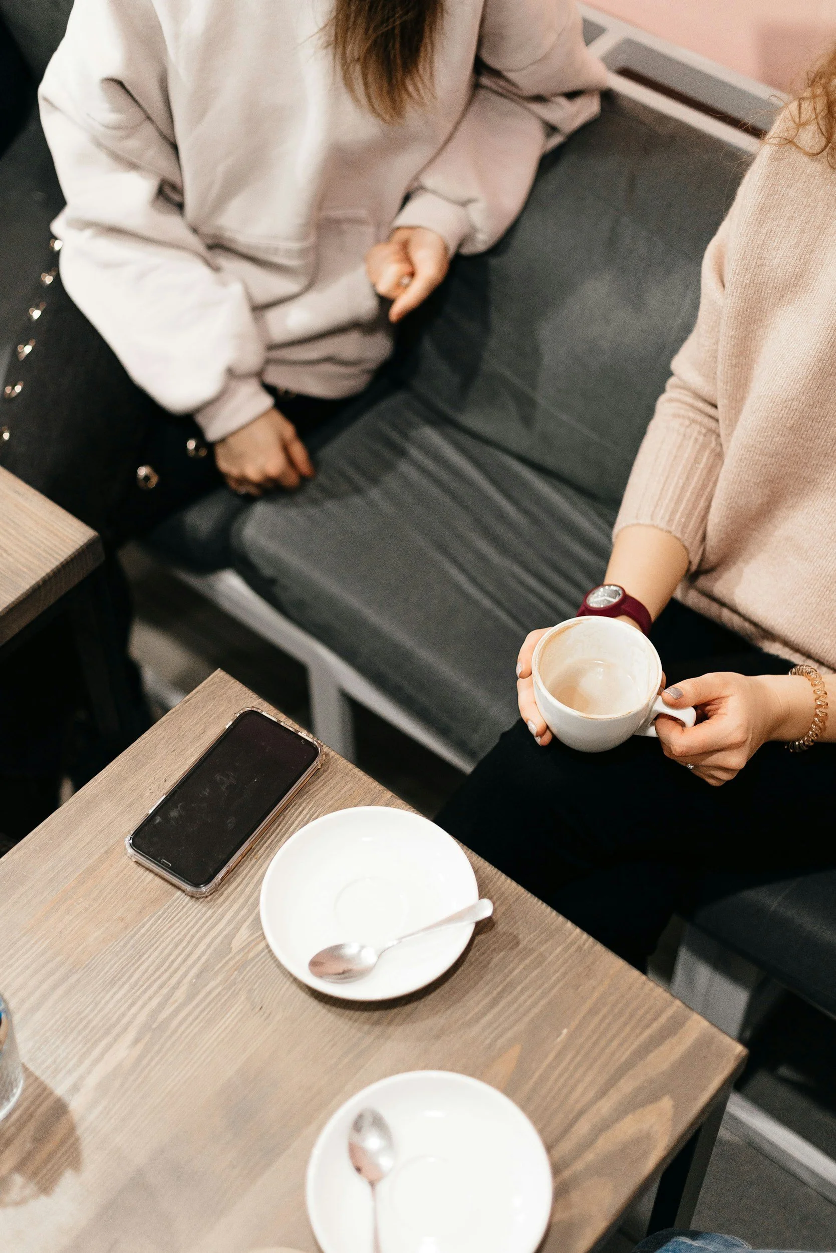 Two women are sitting at a wooden table, one holding a white coffee cup, with empty bowls and a smartphone on the table. One woman is wearing a beige sweater, and the other is wearing a light-colored hoodie. The setting appears to be a casual cafe.