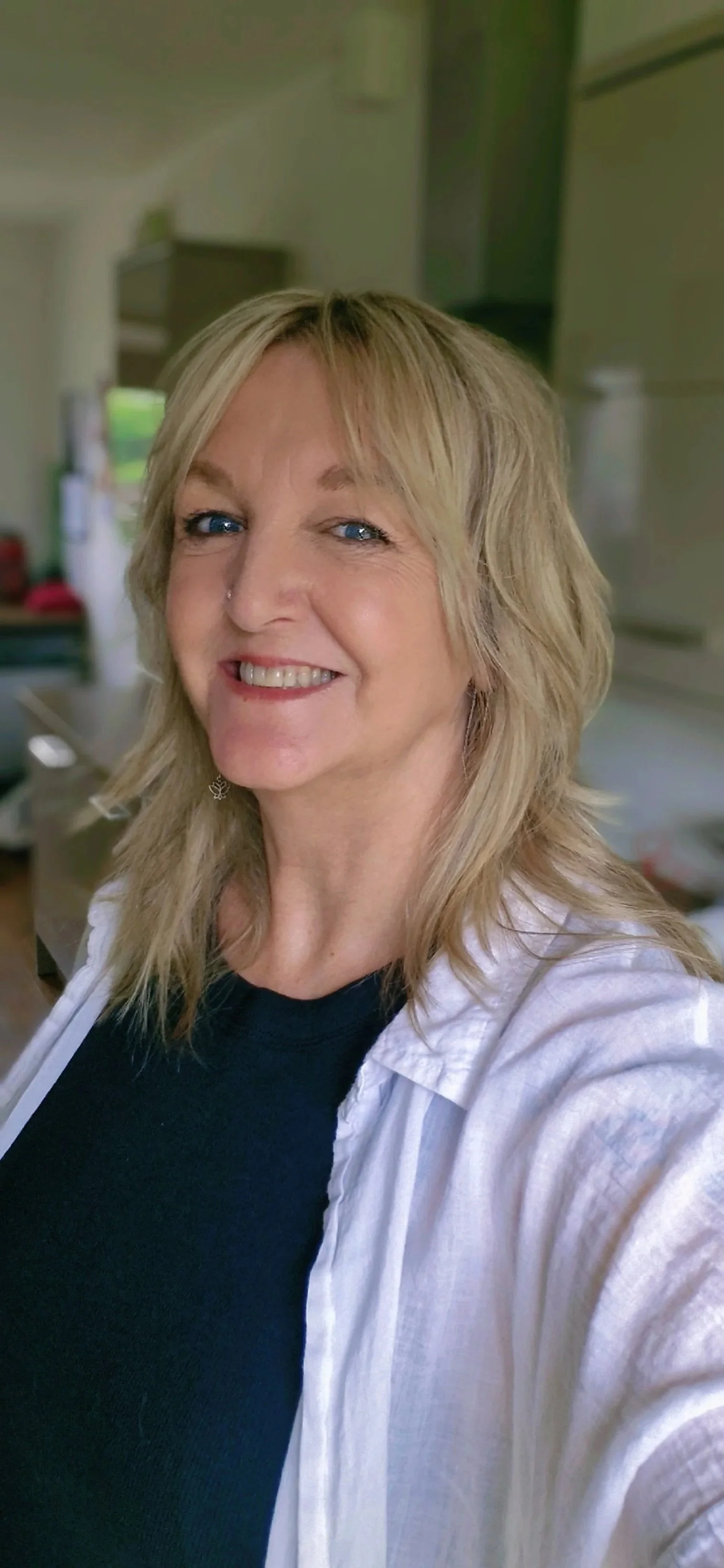 A woman with blonde hair, blue eyes, and a nose piercing smiling in a kitchen.