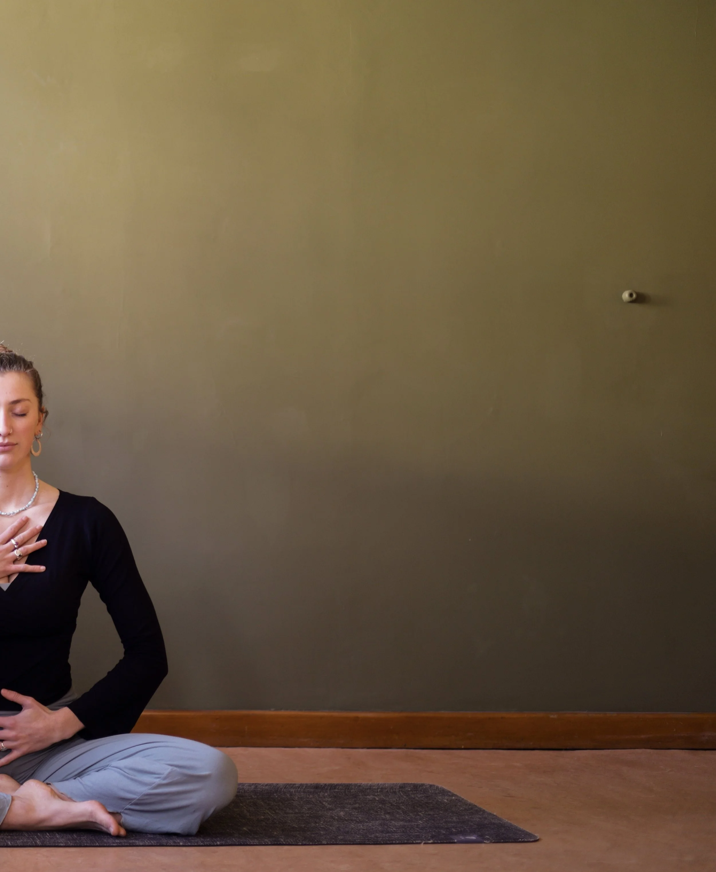 Woman sitting cross-legged on a yoga mat with her hand over her heart, while another person is lying on the mat, practicing yoga or meditation, in a room with a beige wall and wooden floor.