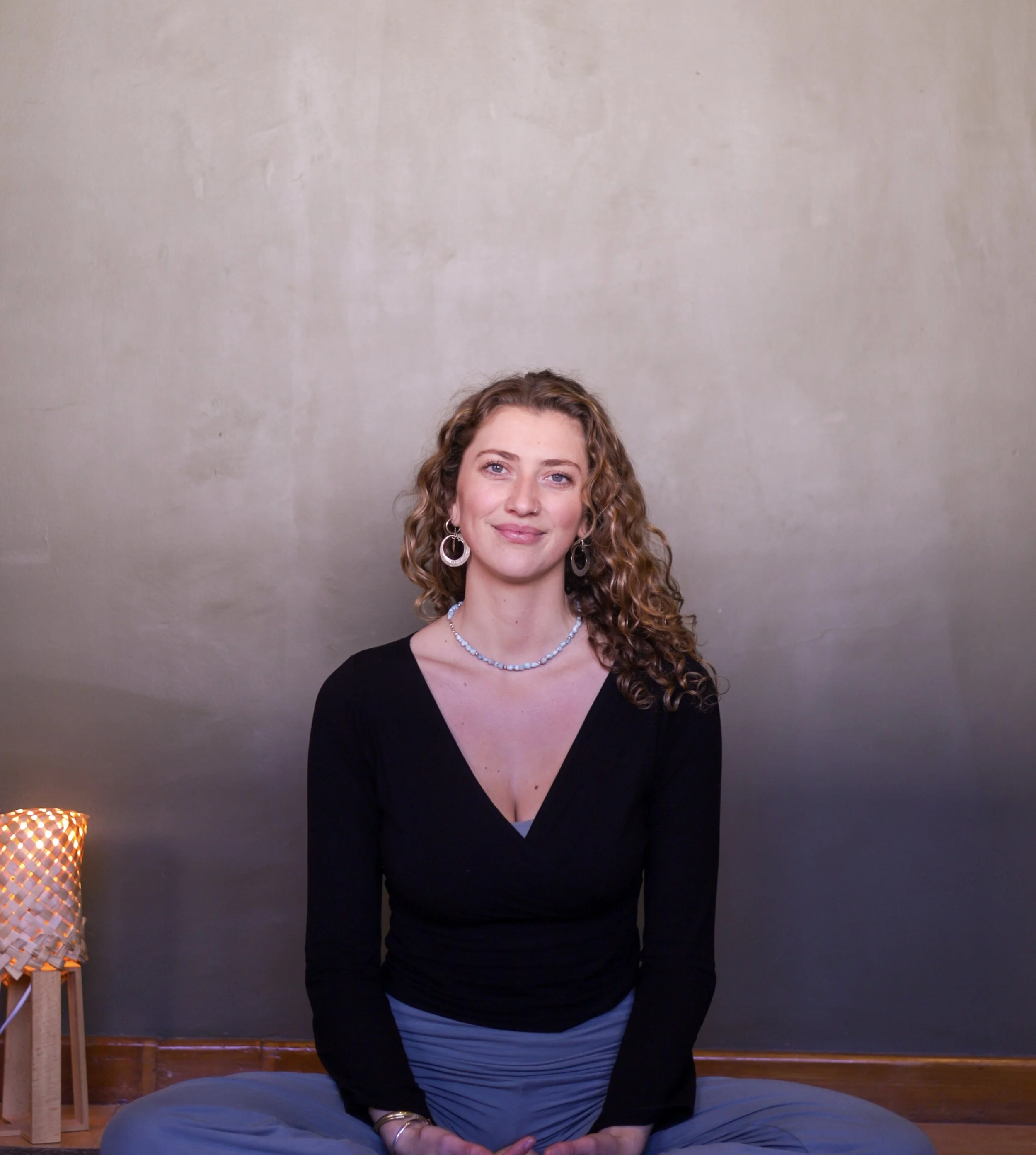 A woman with curly light brown hair and earrings, sitting cross-legged on a blanket, smiling, with a gray wall background and a small lamp on a side table.