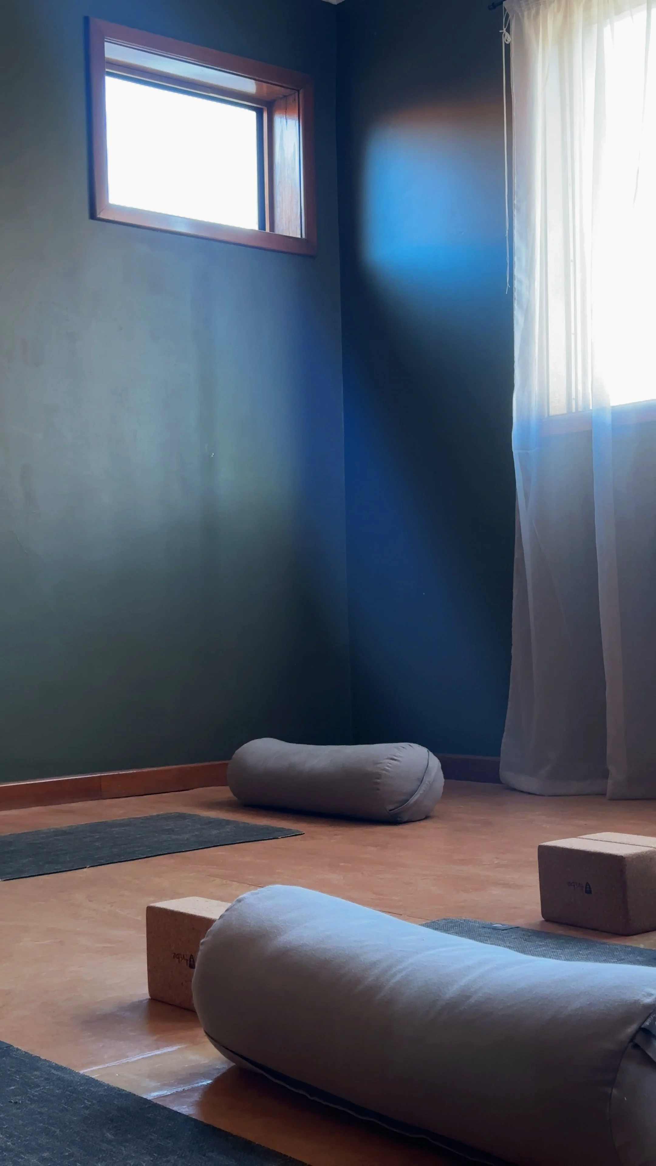 A yoga or meditation studio with blue walls, a window with curtains letting in natural light, and yoga bolsters and blocks on a wooden floor.