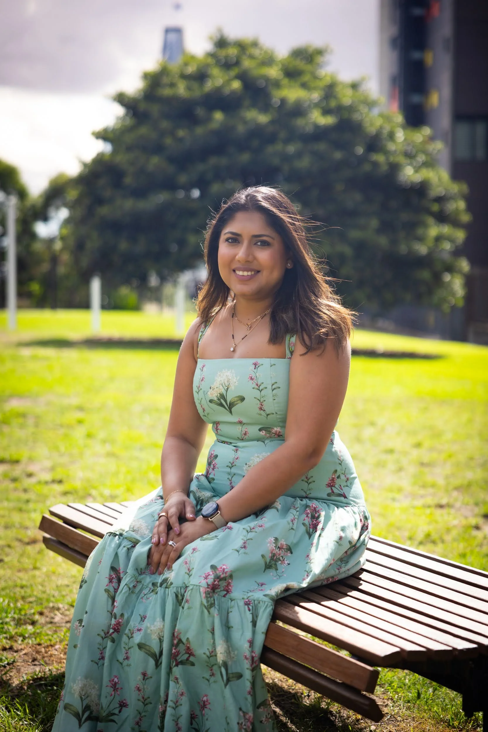 A woman wearing a floral dress sitting on a park bench with green grass and trees in the background under a partly cloudy sky.