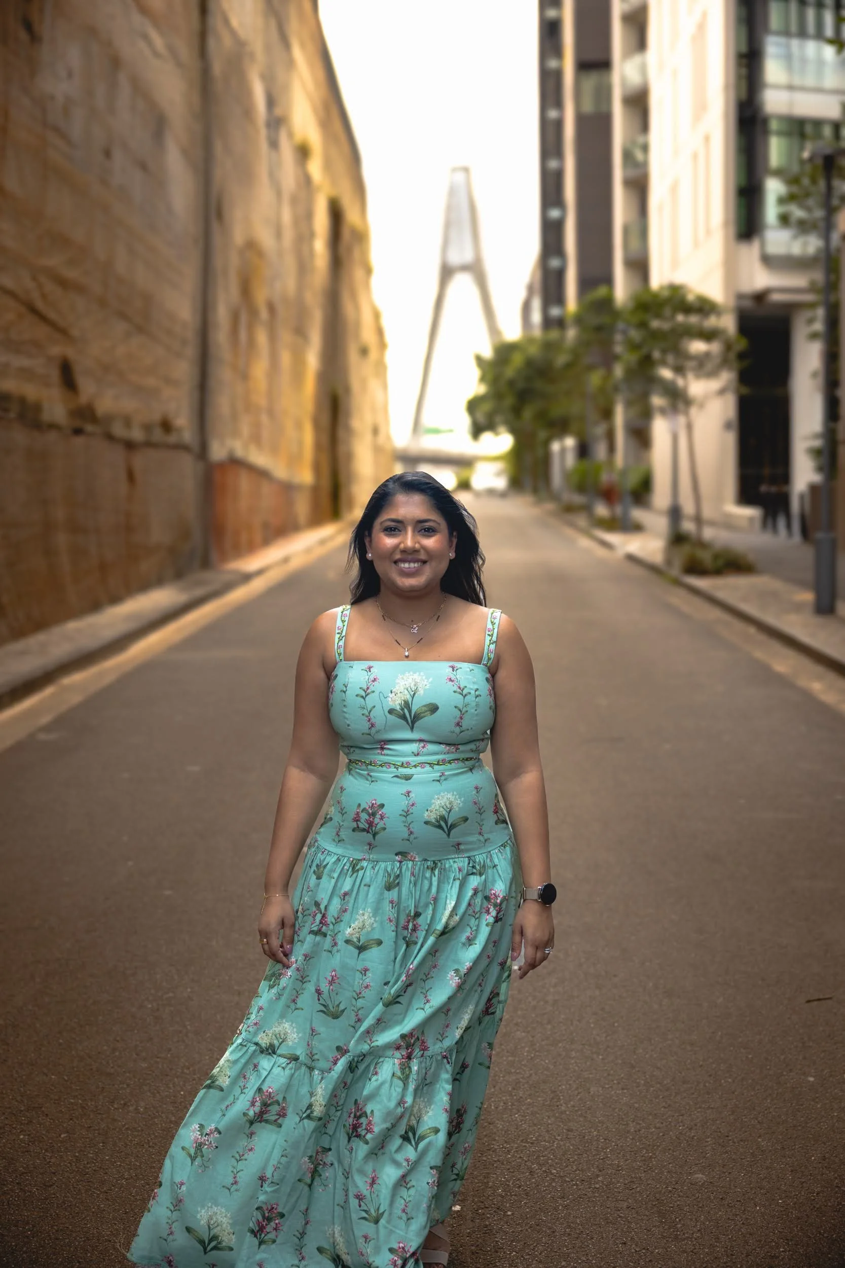 A woman standing on a city street during daylight, wearing a light blue floral dress, smiling, with modern buildings and a suspension bridge in the background.