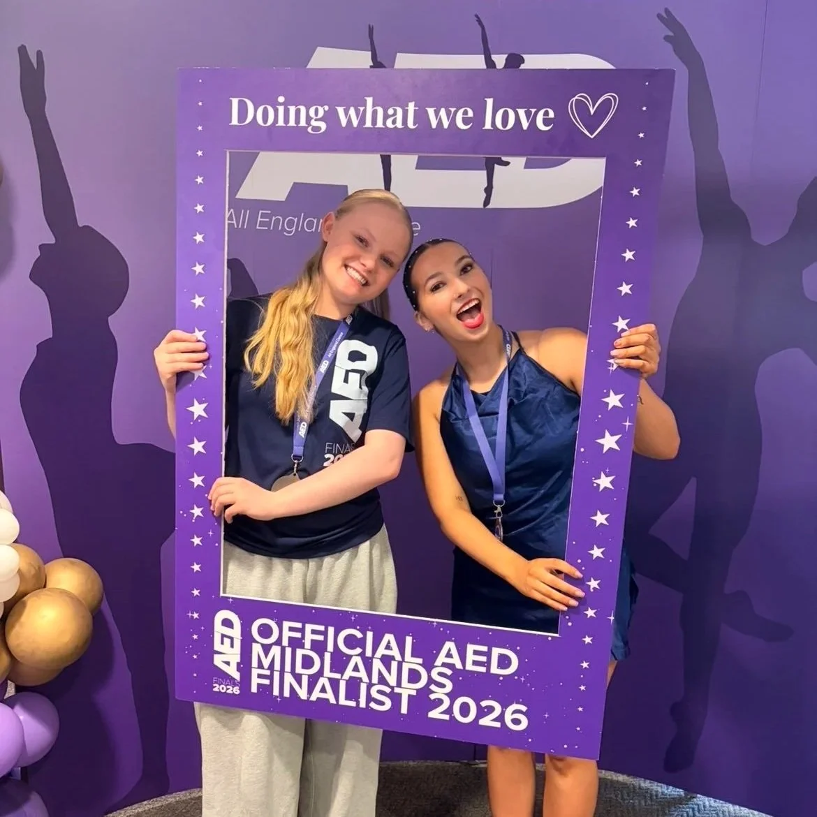 Two young women smiling and holding a large purple frame that reads 'Doing what we love' and 'Official AED Midlands Finalist 2026', standing in front of a purple backdrop.