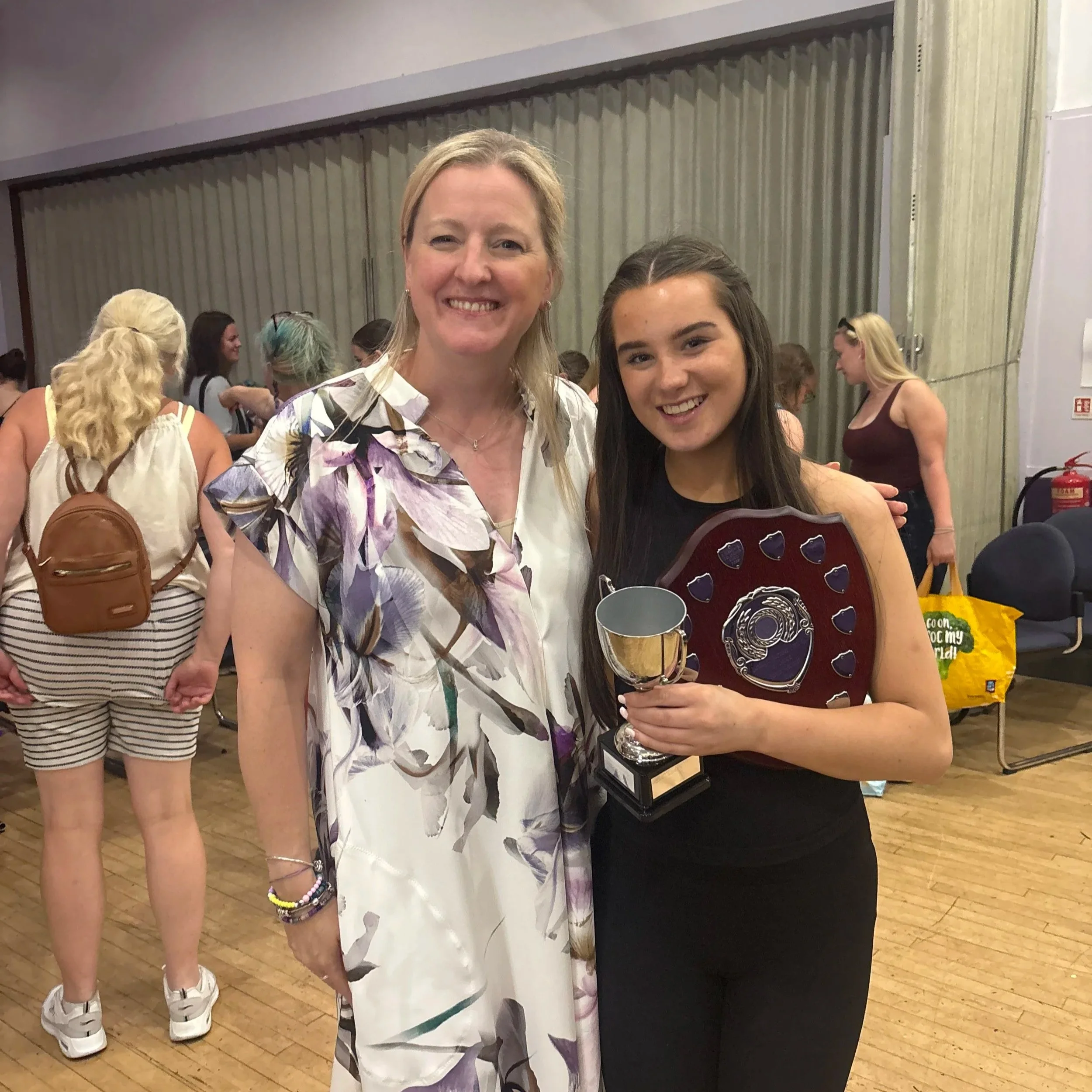 Two women smiling at the camera, one in a floral dress and the other holding a trophy, at an indoor event with people in the background.