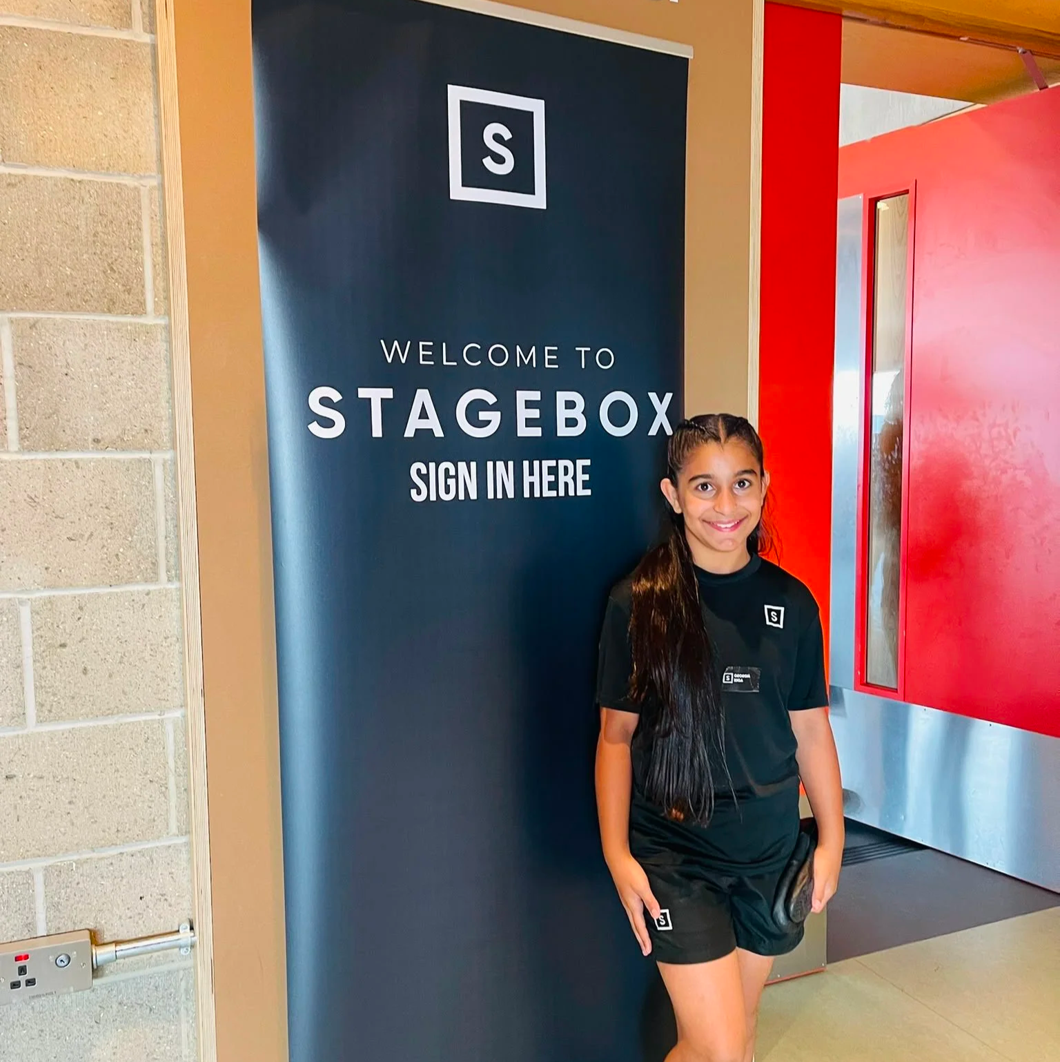 A young girl with long dark hair standing inside a building, smiling at the camera, next to a blue sign that reads 'Welcome to Stagebox Sign In Here'.