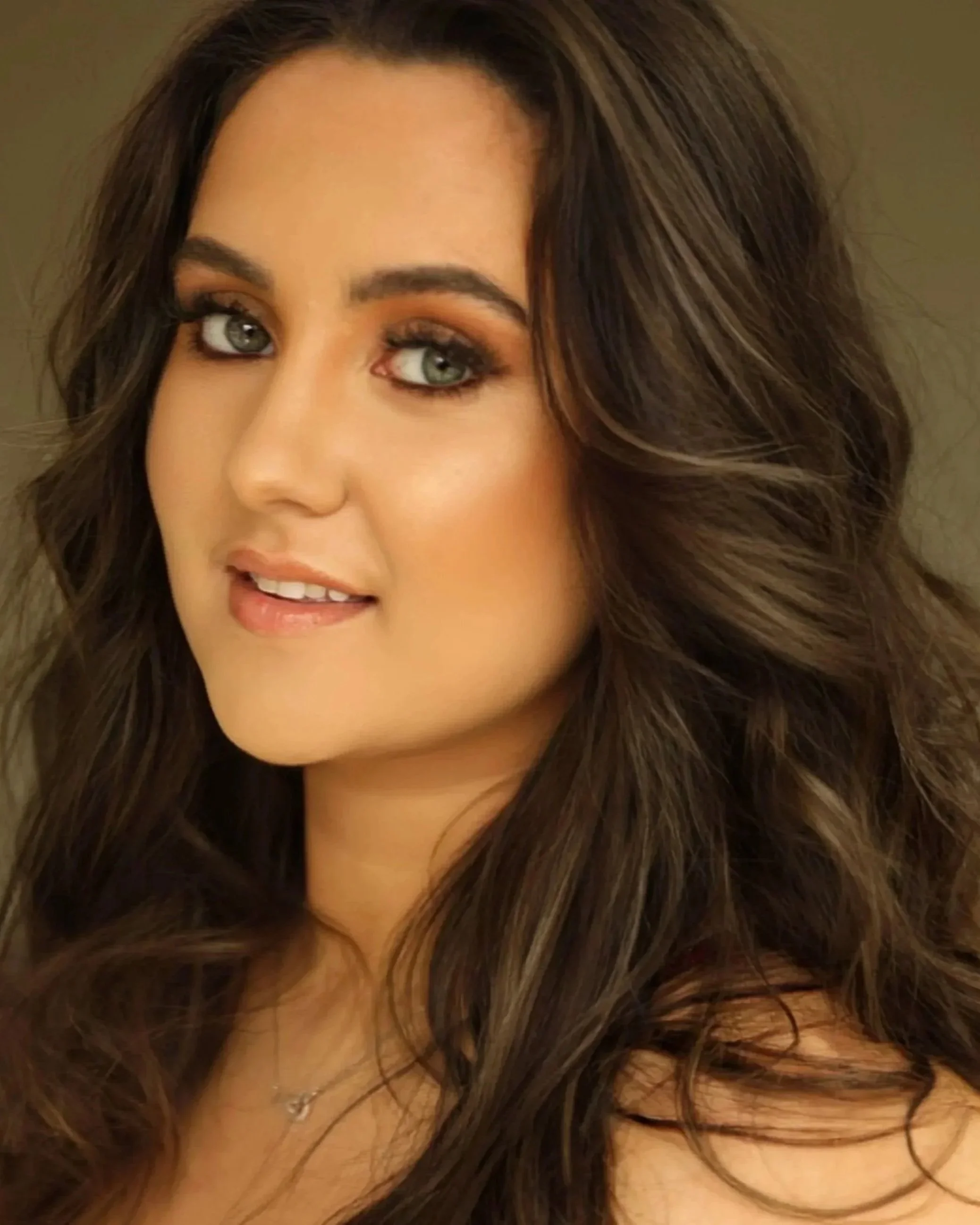 Close-up of a woman's face with long, wavy brown hair, blue eyes, makeup, and a small silver necklace.