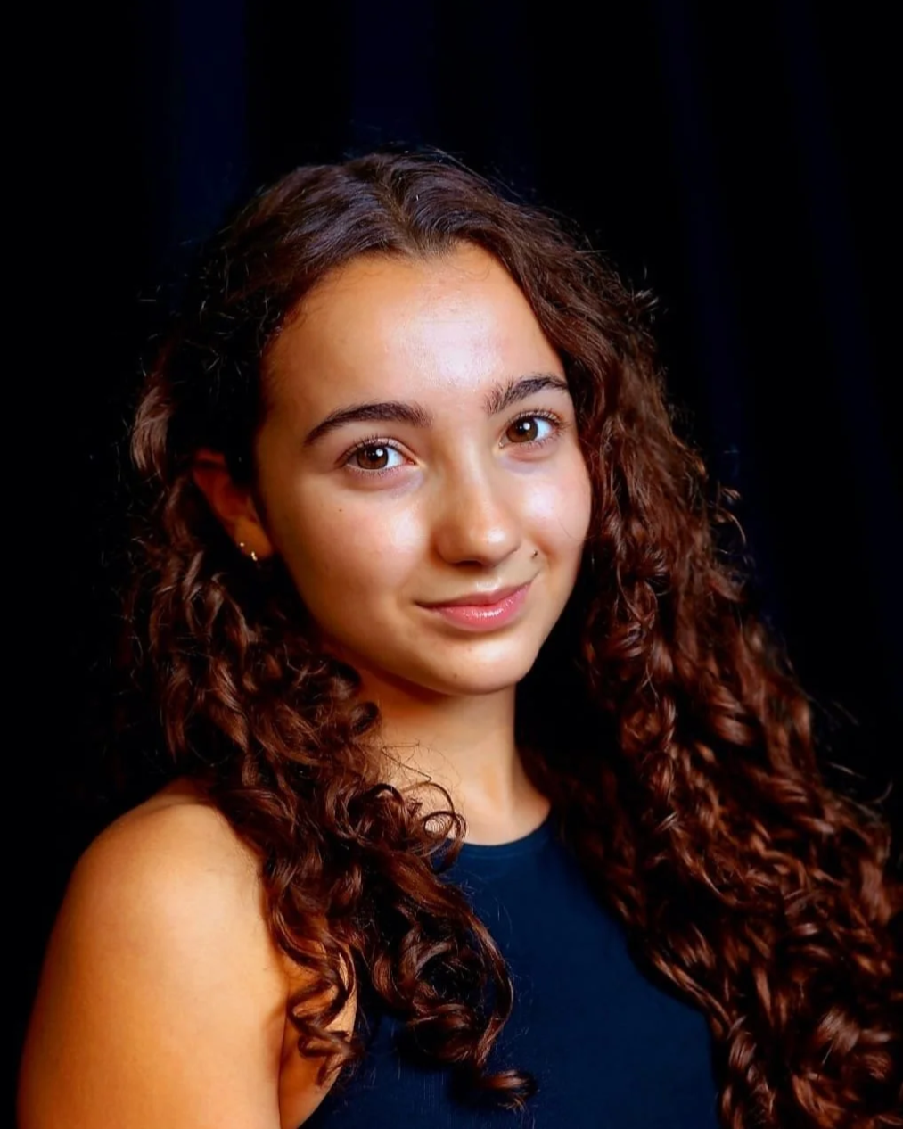 A young woman with long, curly brown hair, wearing a dark sleeveless top, smiling softly against a black background.
