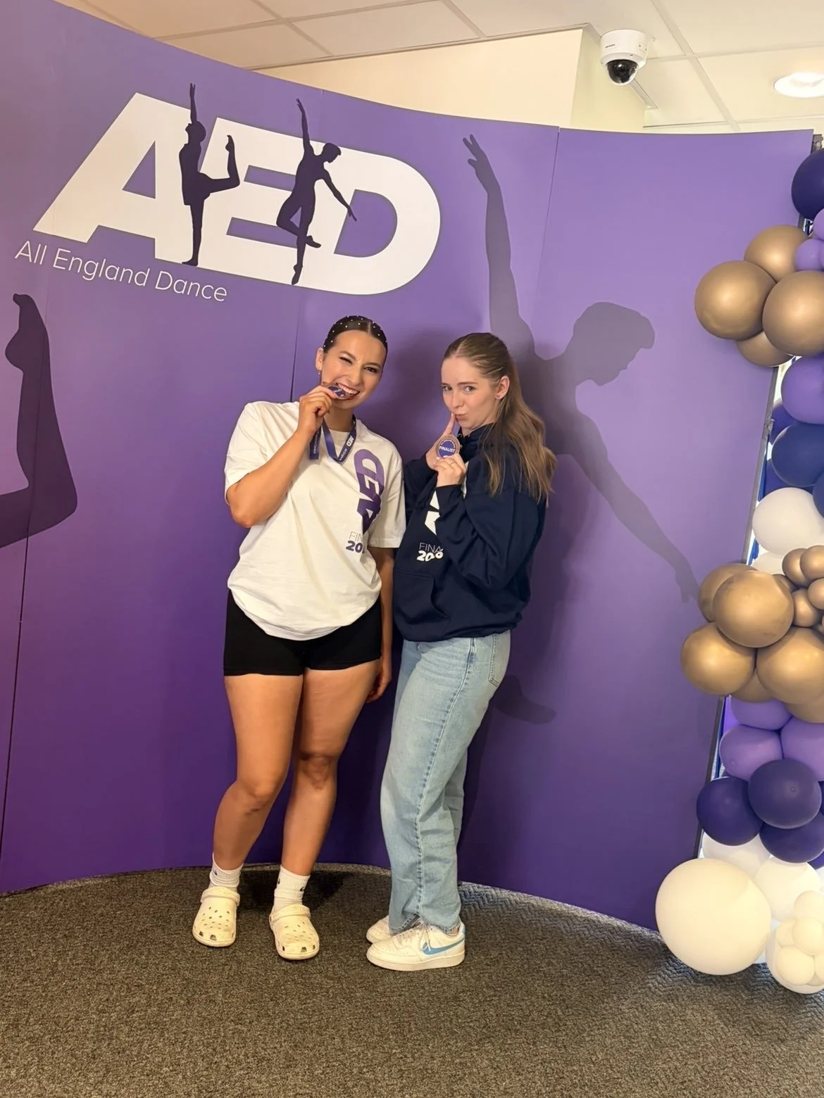 Two young women standing in front of a purple background with the logo 'AED All England Dance,' holding medals and smiling.