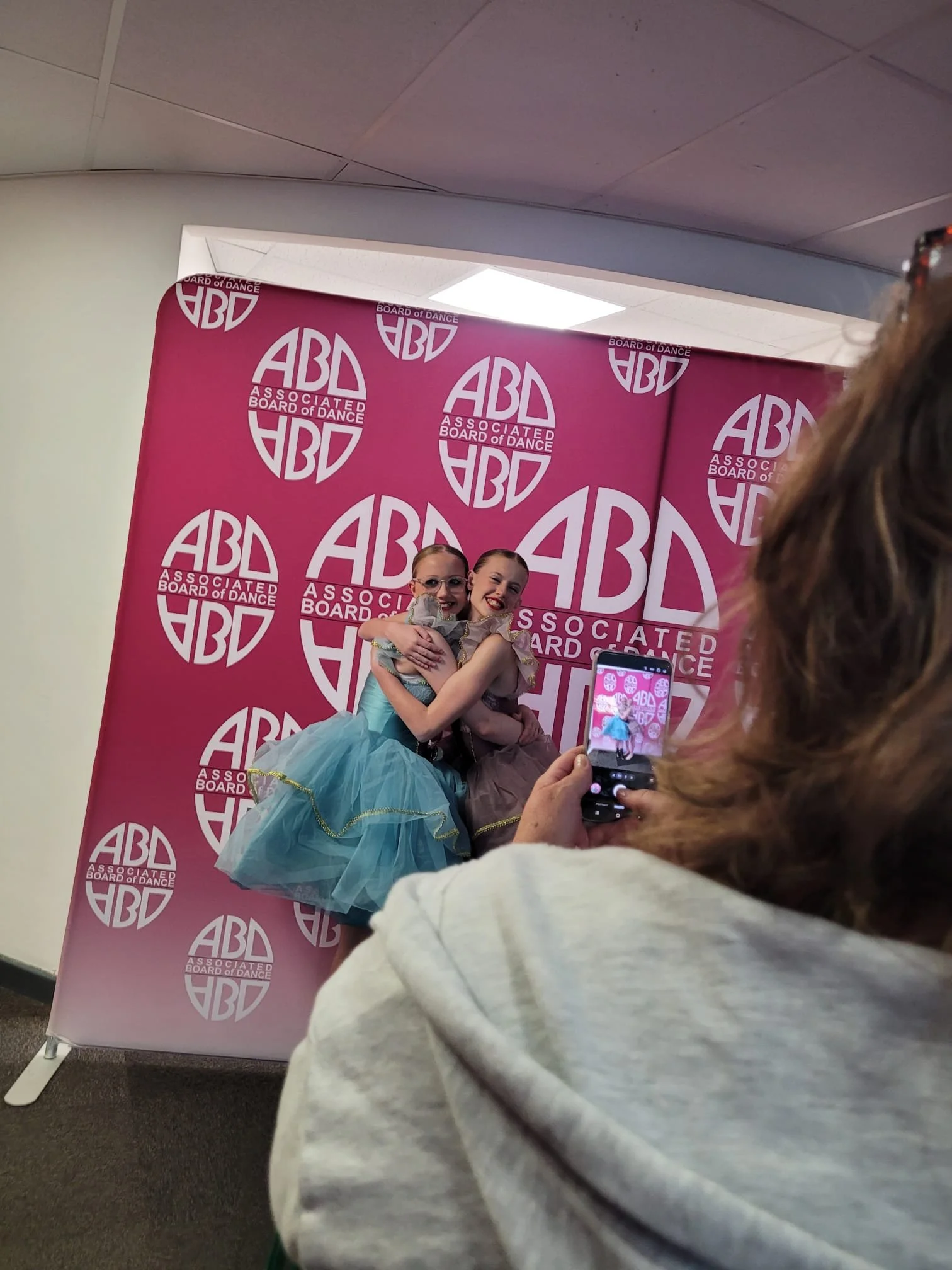 Two young girls in ballet costumes, hugging and smiling, standing in front of a pink backdrop with the 'Associated Board of Dance' logo, during a dance event.