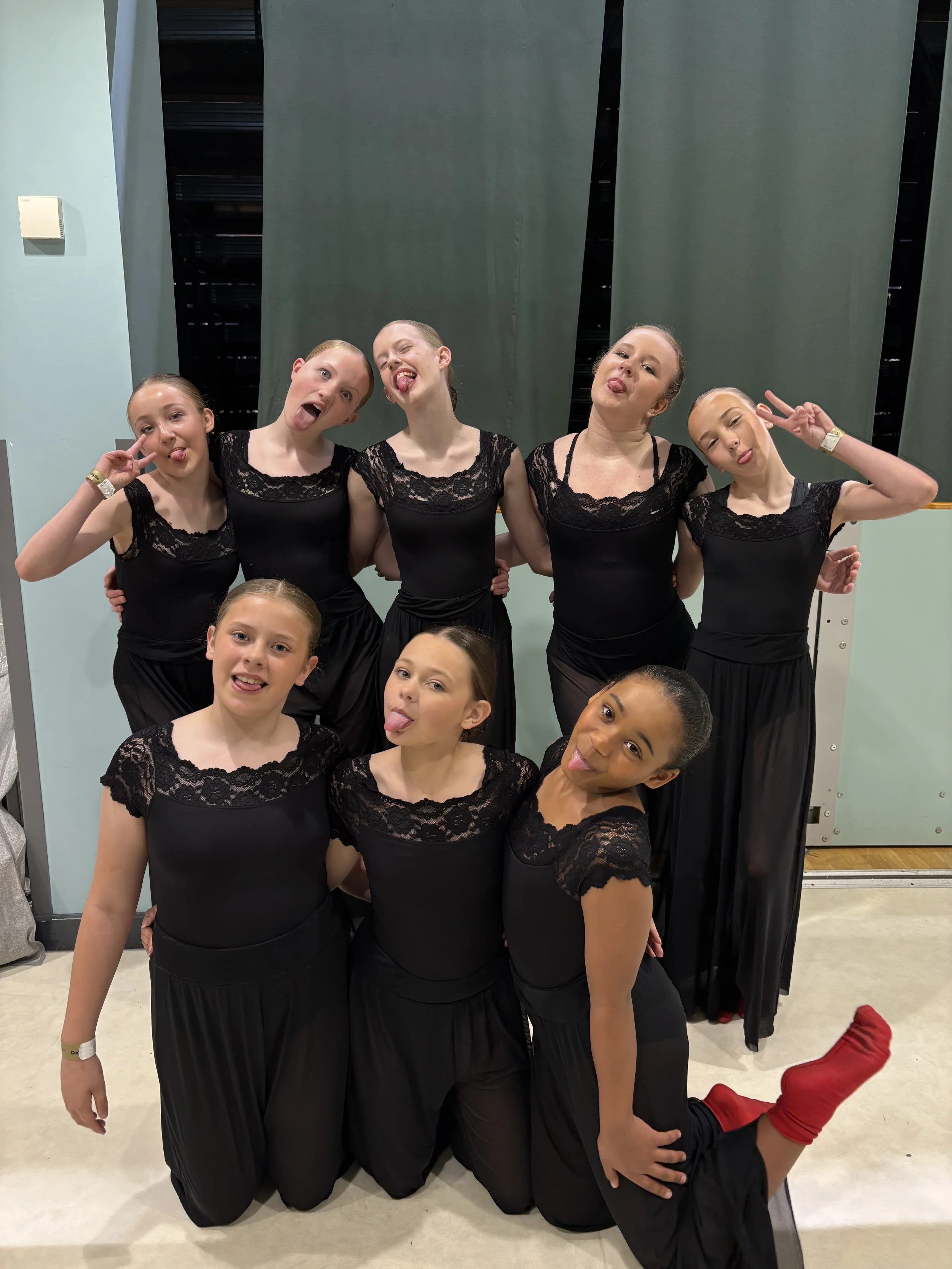 A group of nine young girls dressed in black costumes with lace tops posing for the camera in an indoor setting with green walls and a blue background.