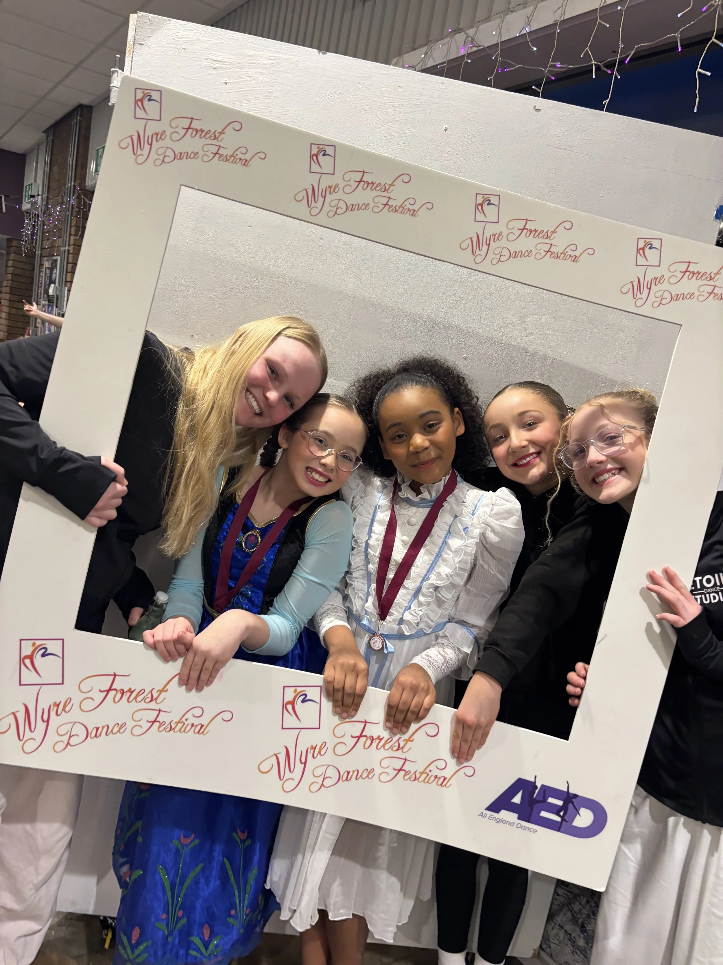 A group of children holding a large frame with the text 'Wyre Forest Dance Festival' printed on it, with some children wearing medals around their necks, at a dance event.