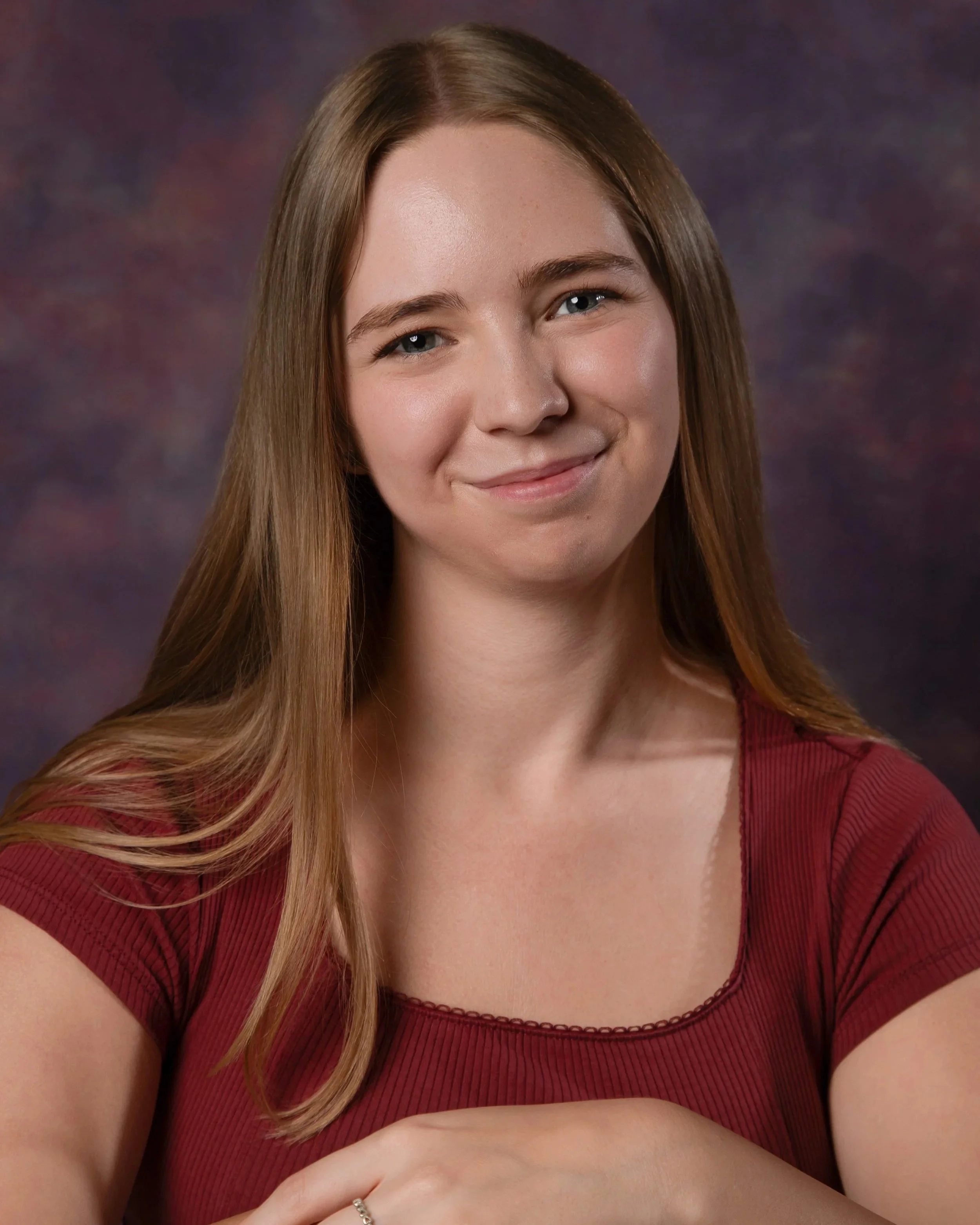 Close-up portrait of a young woman with long, light brown hair, blue eyes, and a gentle smile, wearing a red top with a ribbed texture, against a dark, blurred background.