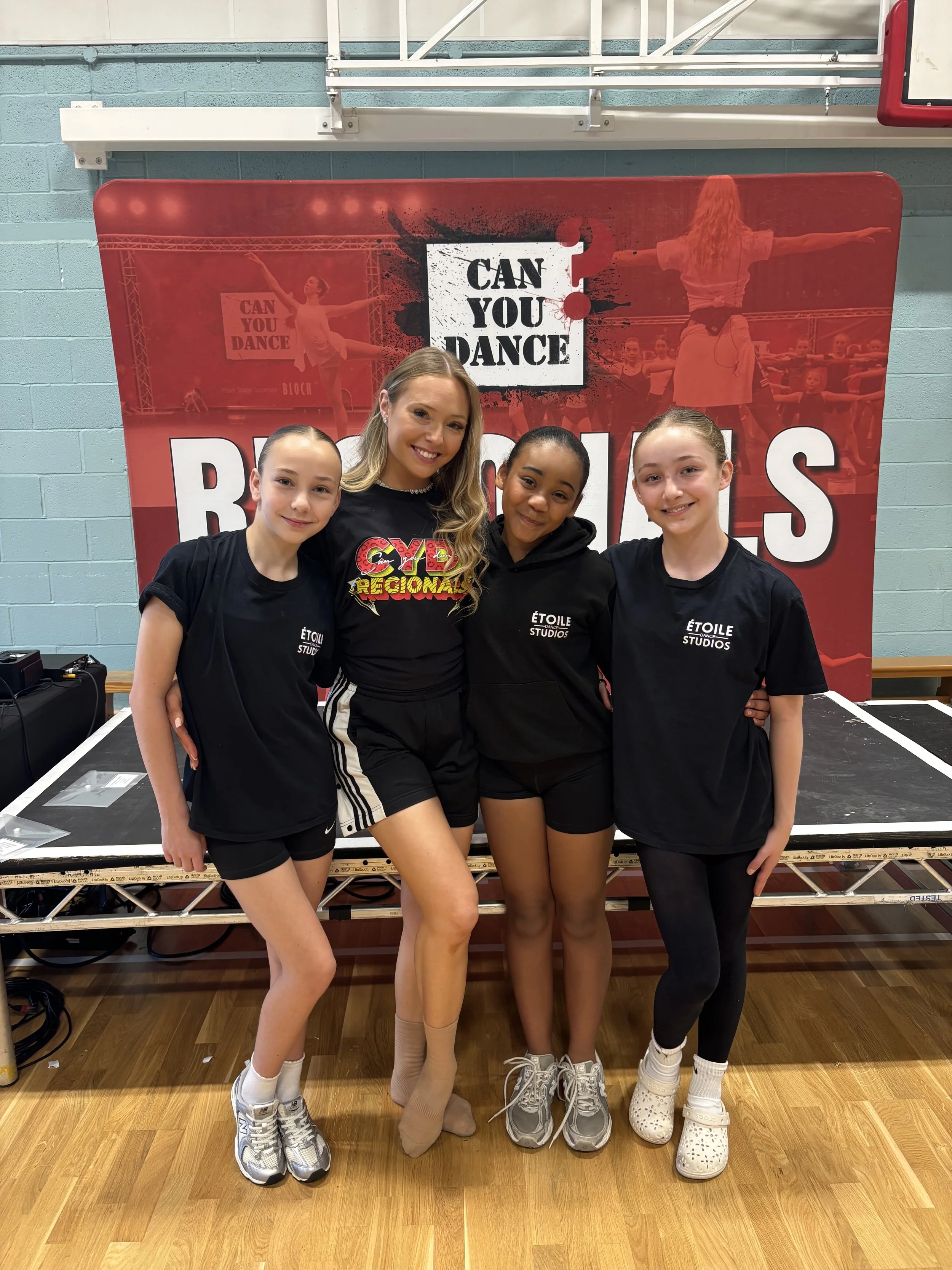 Four young female dancers standing together in front of a red dance studio banner, smiling at the camera, with a professional dance studio backdrop.