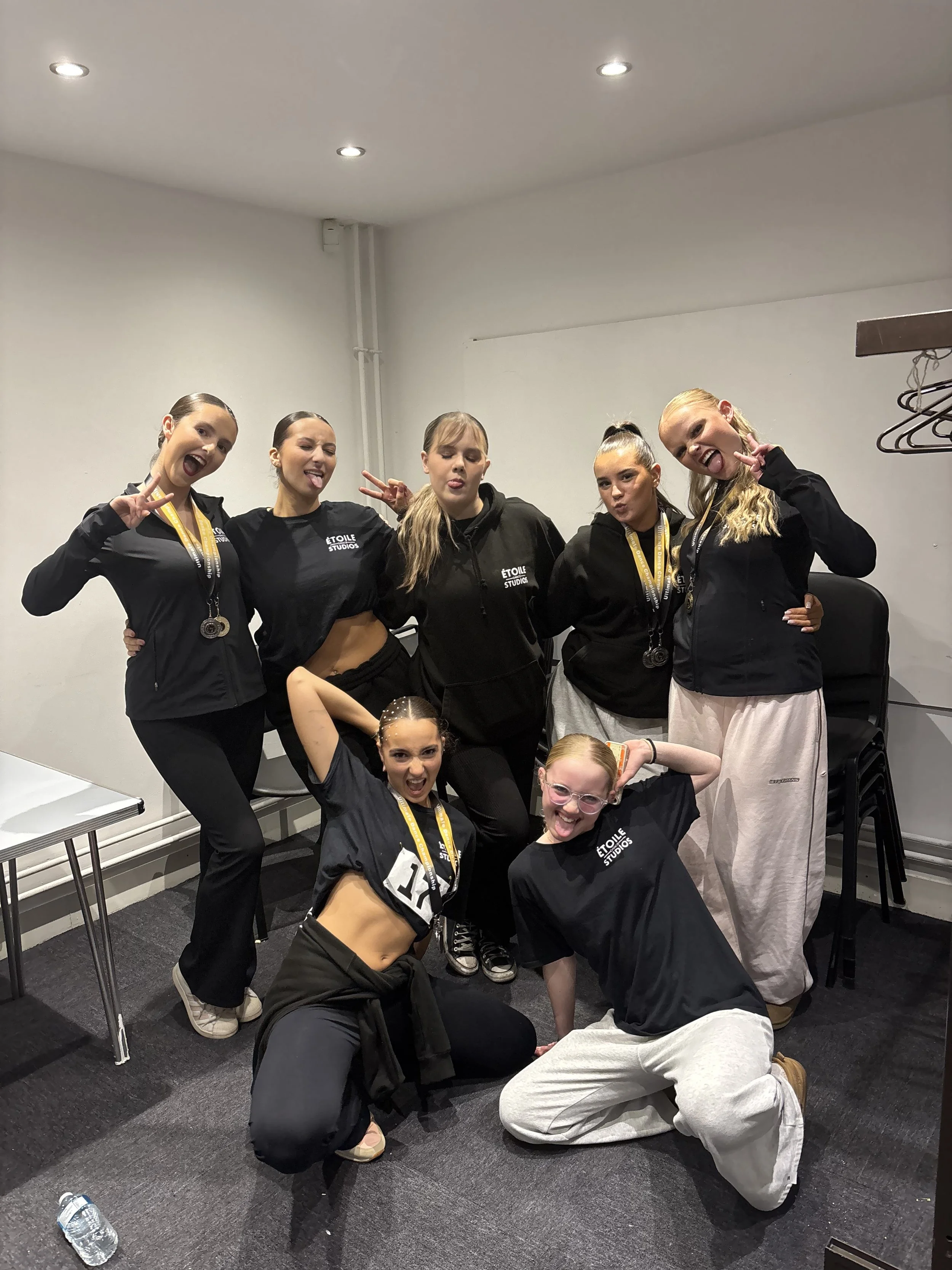 Seven young women, some with medals around their necks, posing together indoors. They are wearing black jackets or sweatshirts with 'Etoile Dance Studios' printed on them.