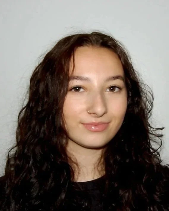 A young woman with long curly brown hair, and a nose piercing, smiling softly against a plain white wall background.