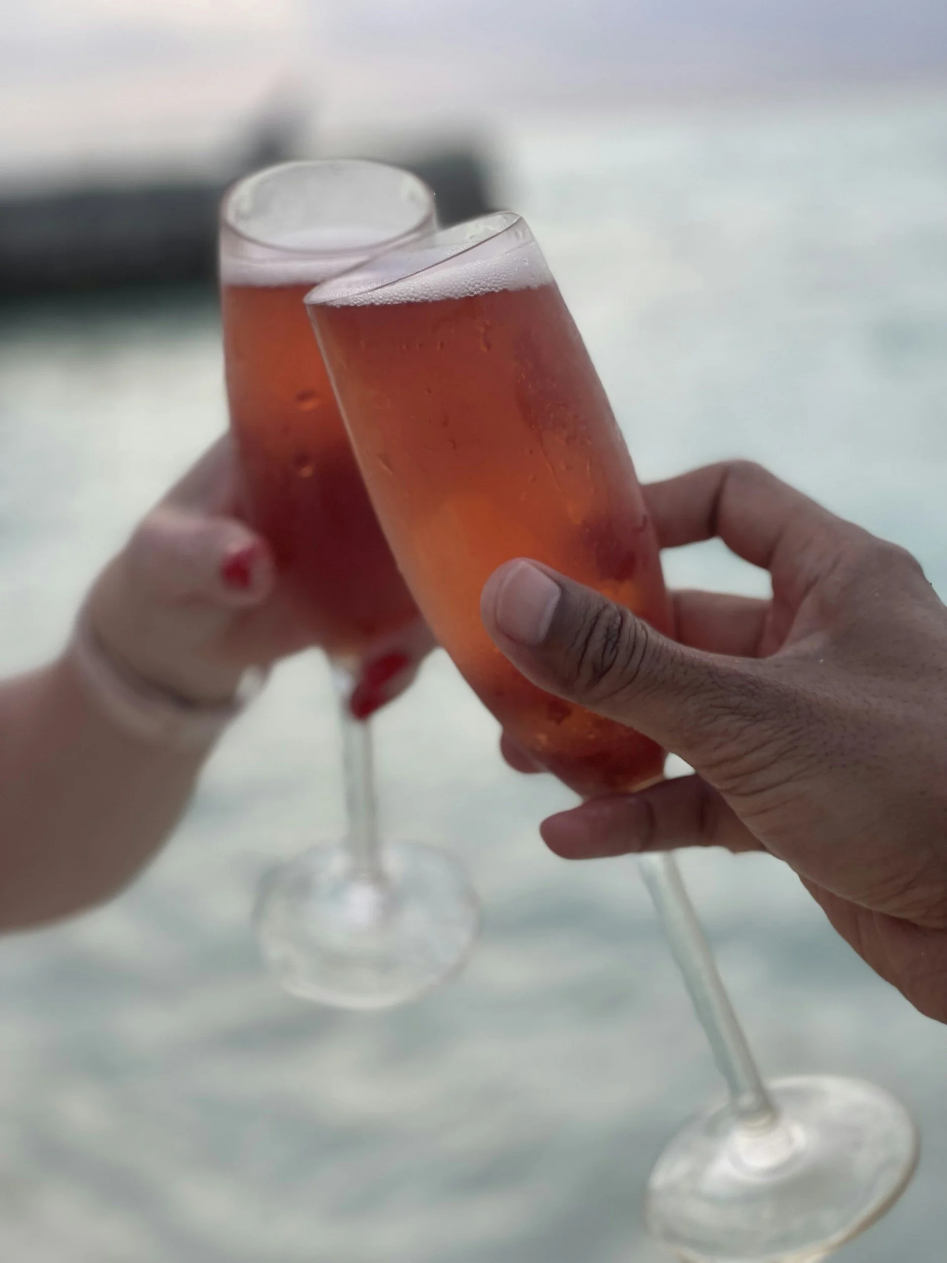 Two hands holding glasses of pink bubbly beverage for a toast, with water droplets on the glasses, against a blurred outdoor background.