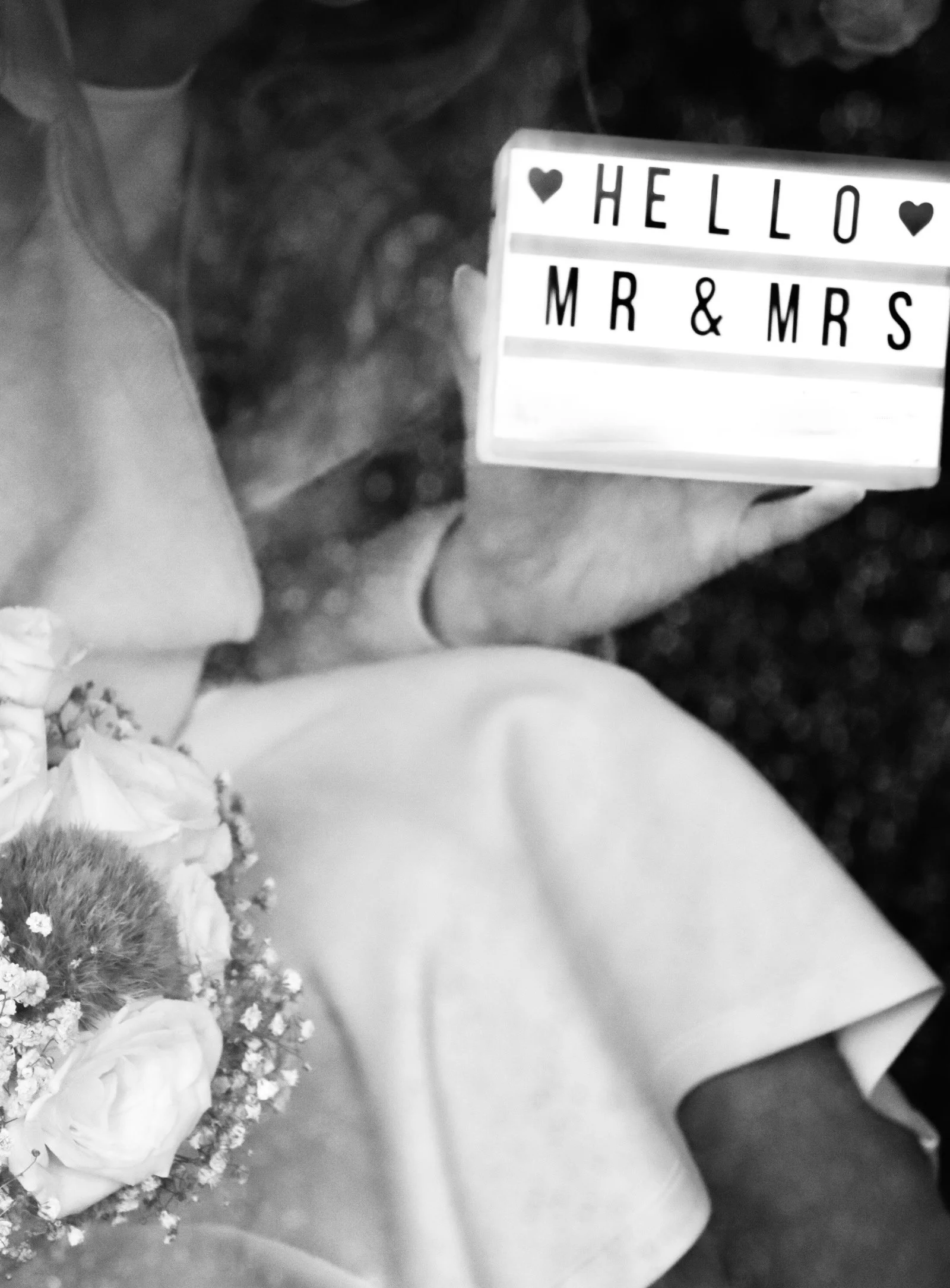 Schwarz-Weiß-Foto eines Hochzeitspaares mit Blumenstrauß und Schild „Hello Mr & Mrs“.