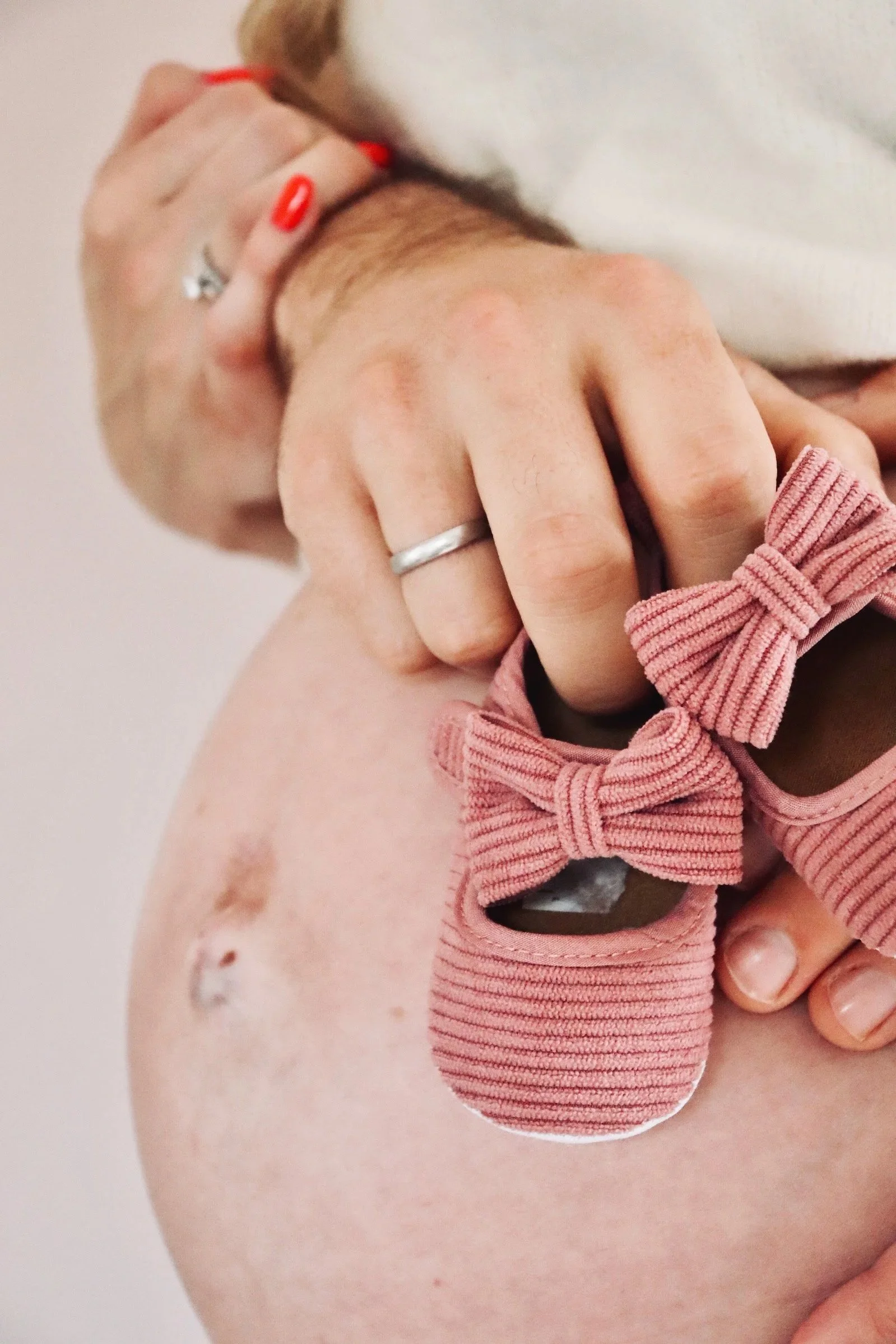 Nahaufnahme eines Babybauches davor ein rosa gestrickter Babyschuh mit Schleife, der von der Hand einer Person mit rotem Nagellack gehalten wird.