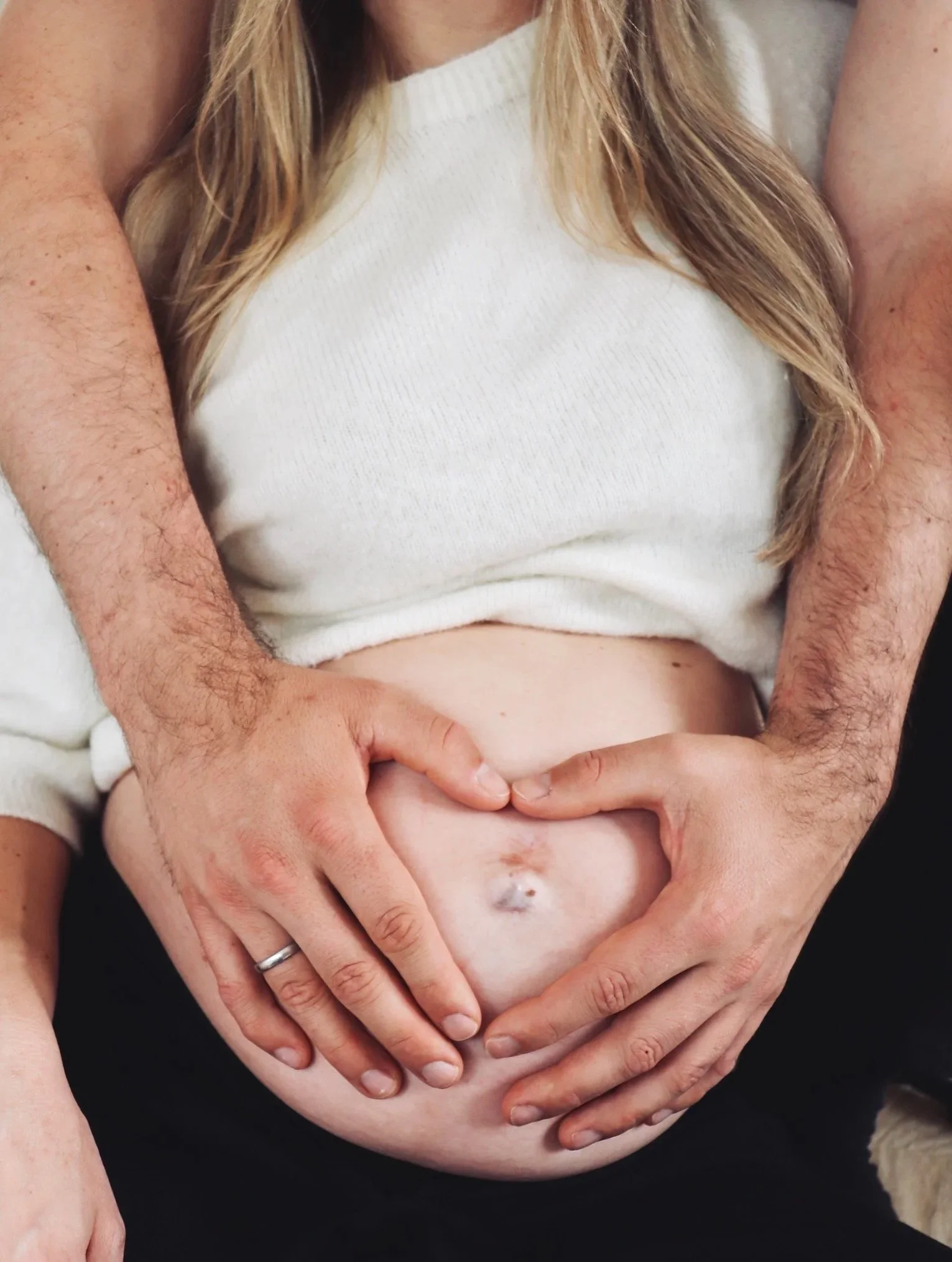 Schwangere Person im weißen Pullover, deren Babybauch von zwei Händen behutsam gehalten wird, welche auf dem Bauch ein Herz formen