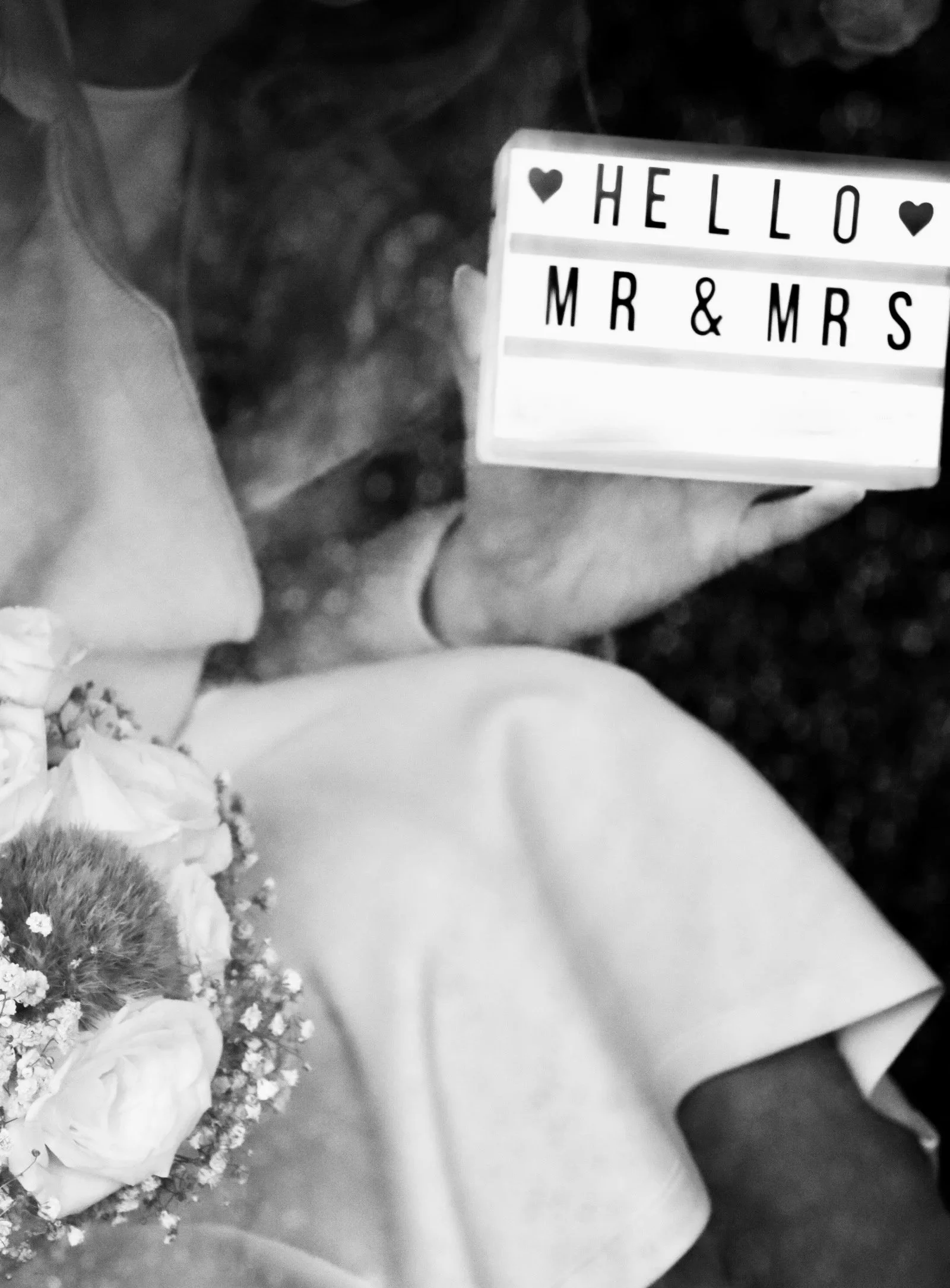Schwarz-Weiß-Foto eines Hochzeitspaares mit Blumenstrauß neben einem Schild mit der Aufschrift „HELLO MR & MRS“.