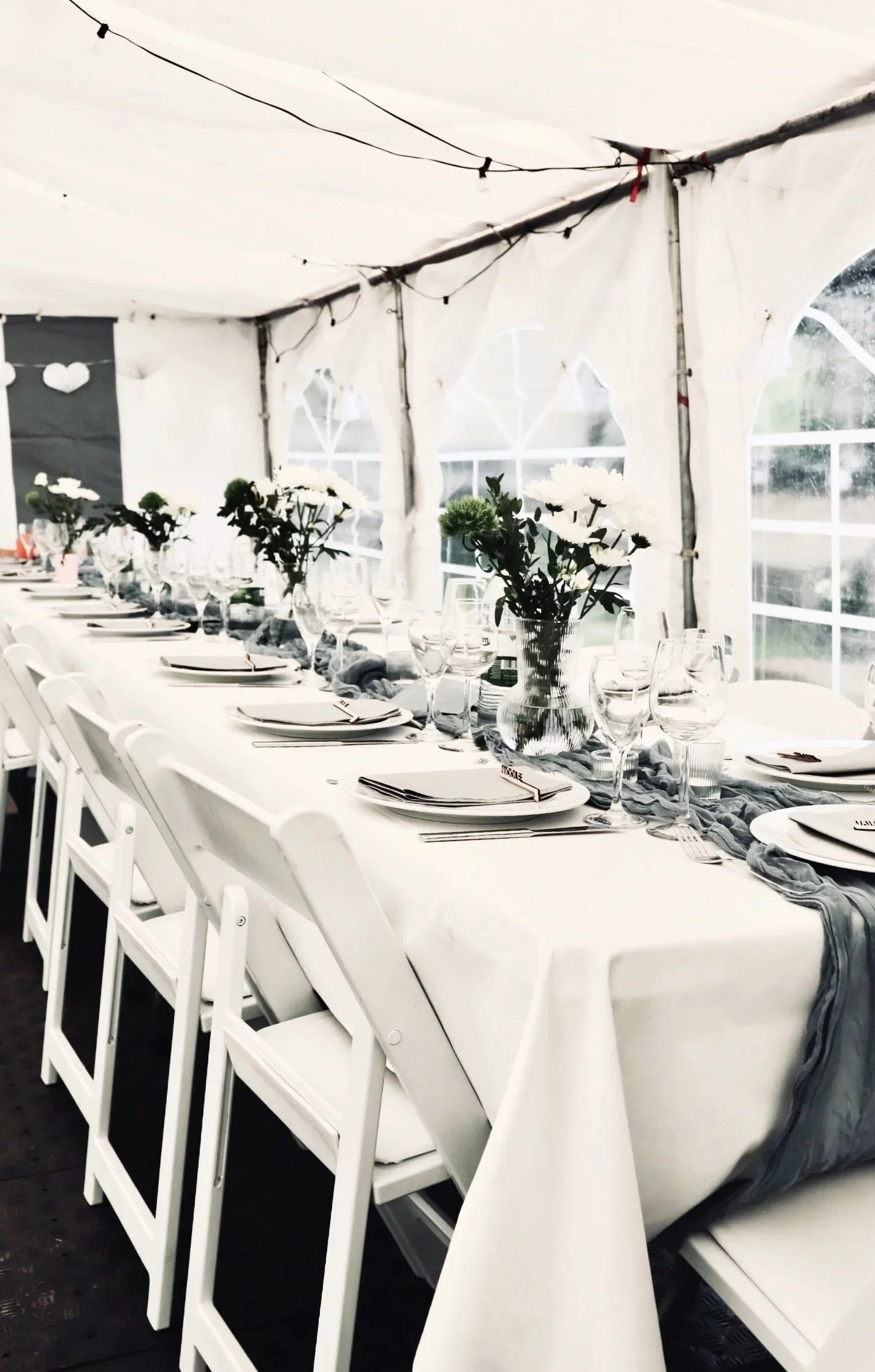 Festlich gedeckte Hochzeitstafel im weißen Festzelt mit Blumenarrangements, Glasgeschirr und weißen Klappstühlen.